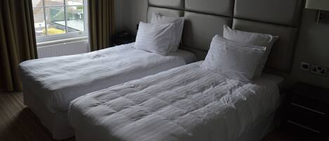 A hotel room with two beds, a window with curtains, a nightstand, and a wall-mounted lamp.