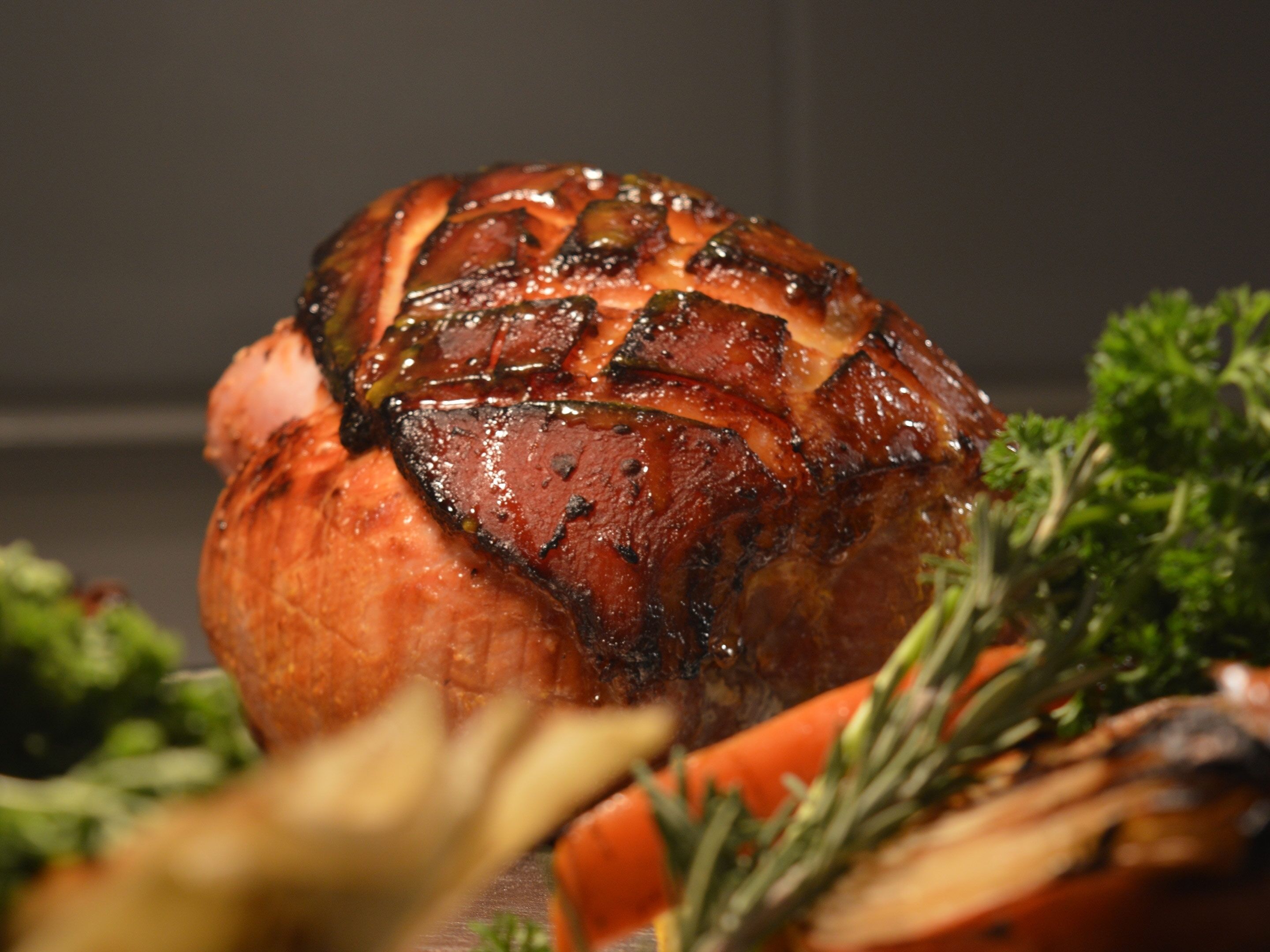 Un jamón asado con corteza brillante, rodeado de verduras como zanahorias y perejil.