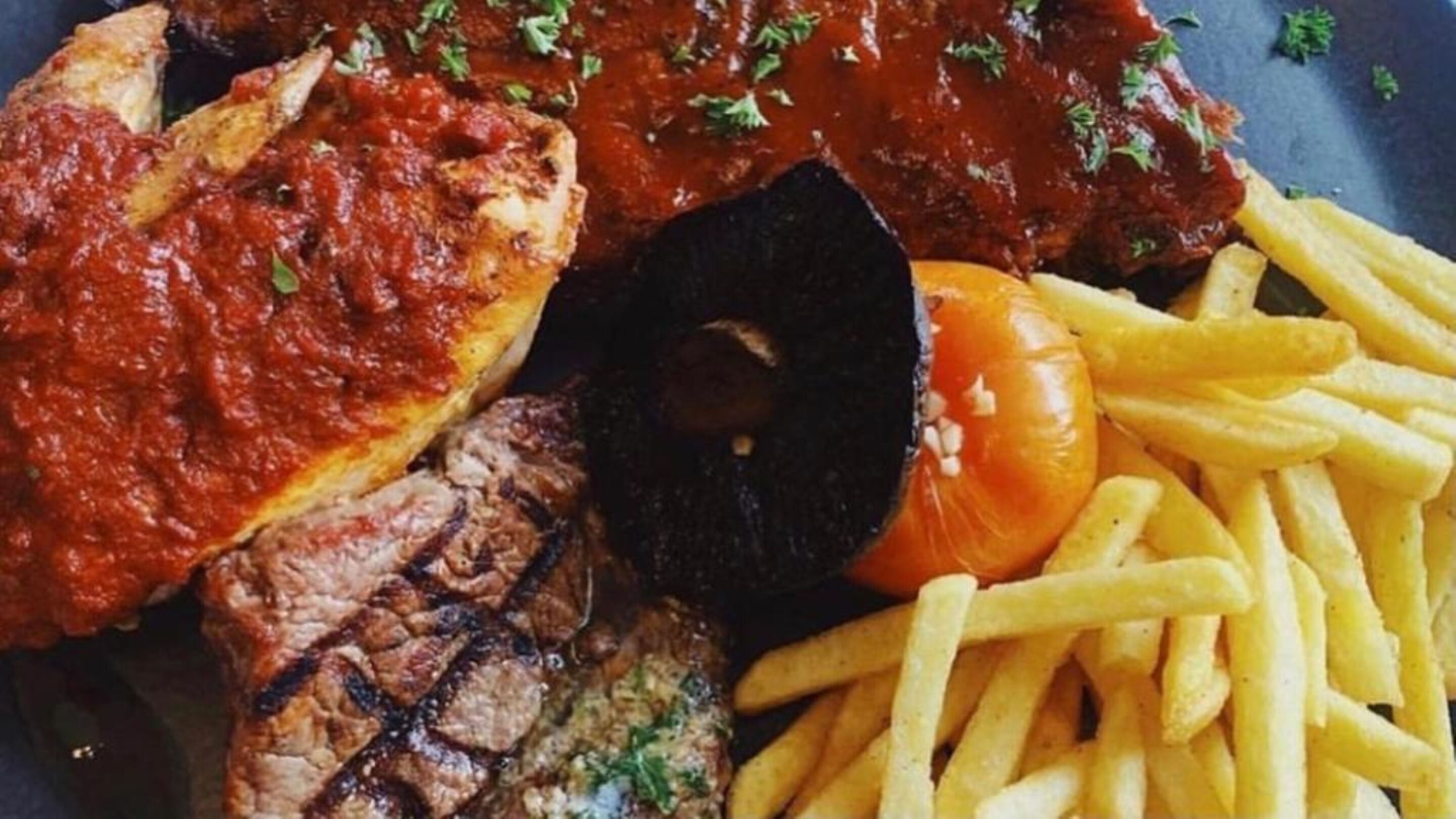 A plate with grilled meat, a slice of pizza, a grilled tomato, and French fries.
