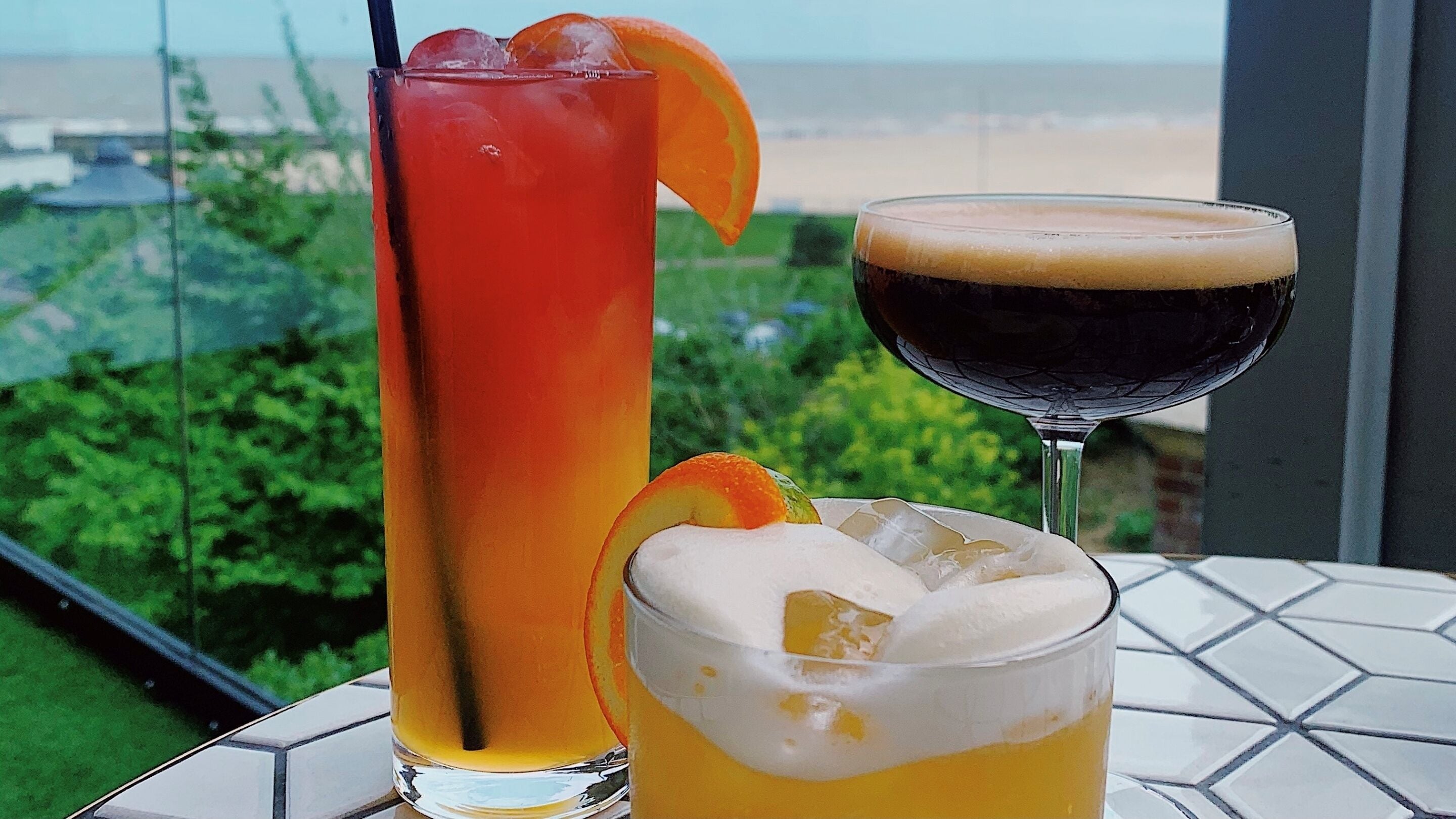 Two drinks on a table with a view of greenery and a building in the background.