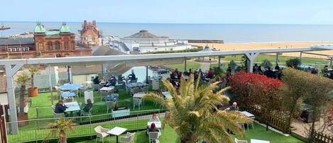 An outdoor patio with tables and chairs, overlooking a beach and a historic building.