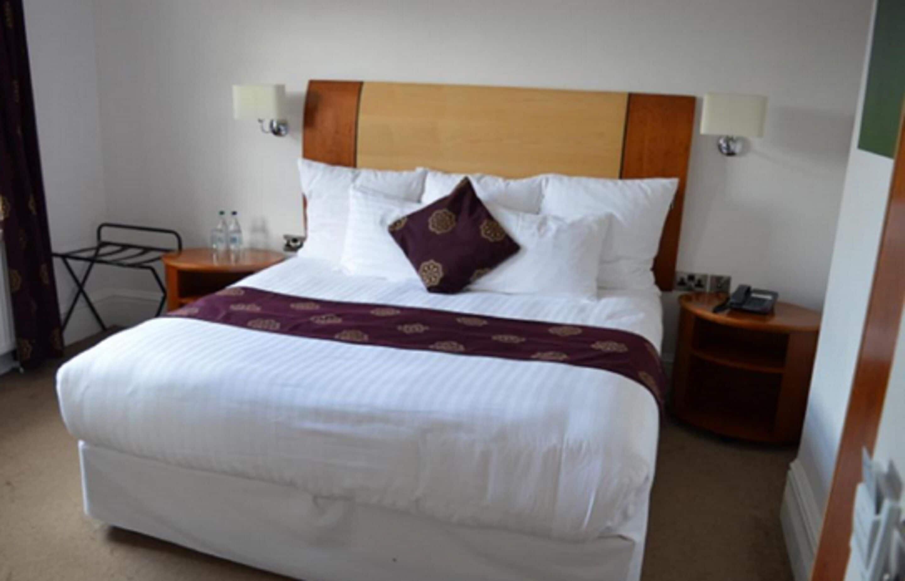 A neatly made bed with a wooden headboard, white bedding, and a decorative pillow. There are two bedside tables, one with a lamp and the other with a phone. A small table with water bottles is visible in the corner.