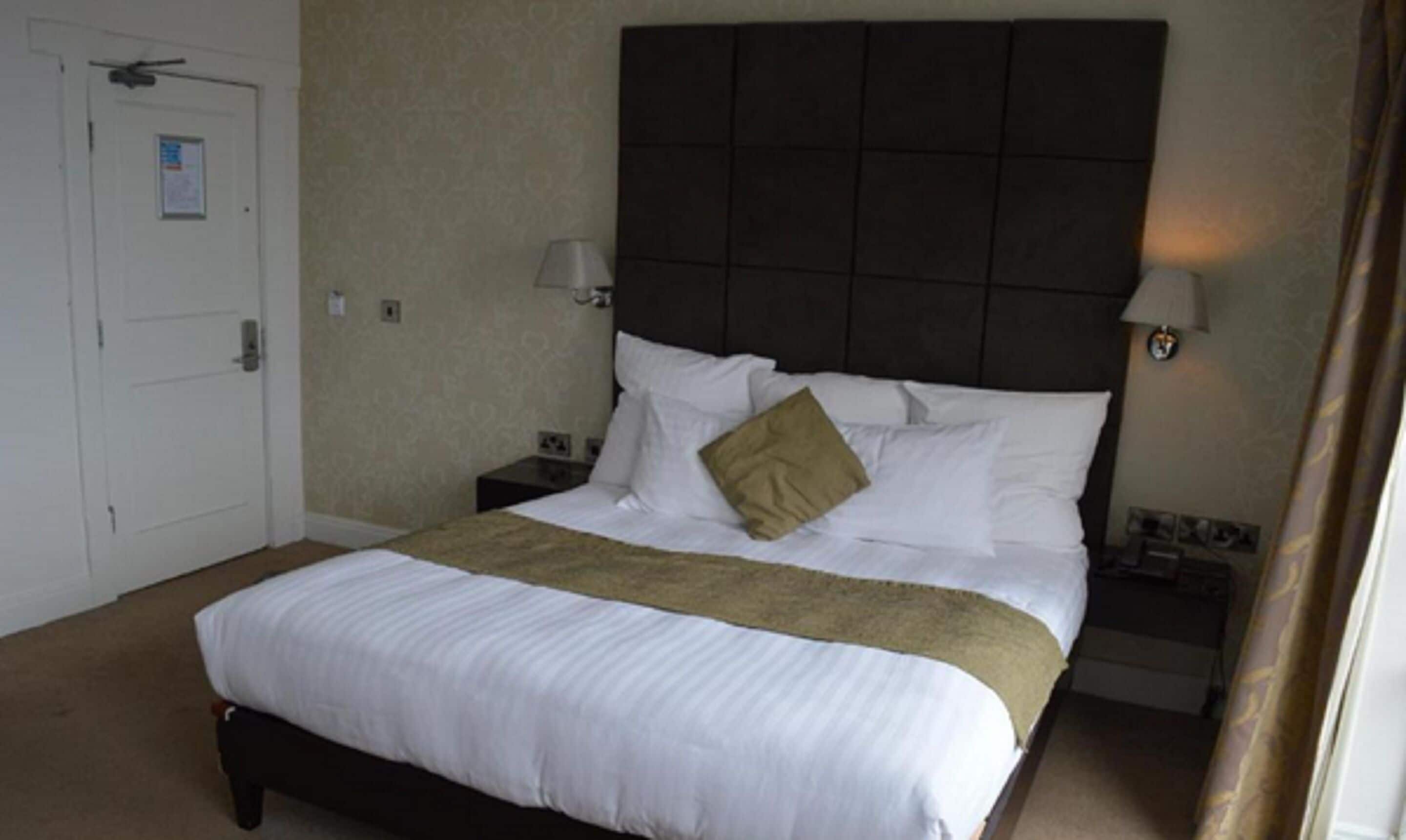 A hotel room with a large bed, two bedside tables, a dark headboard, and a door with a sign.