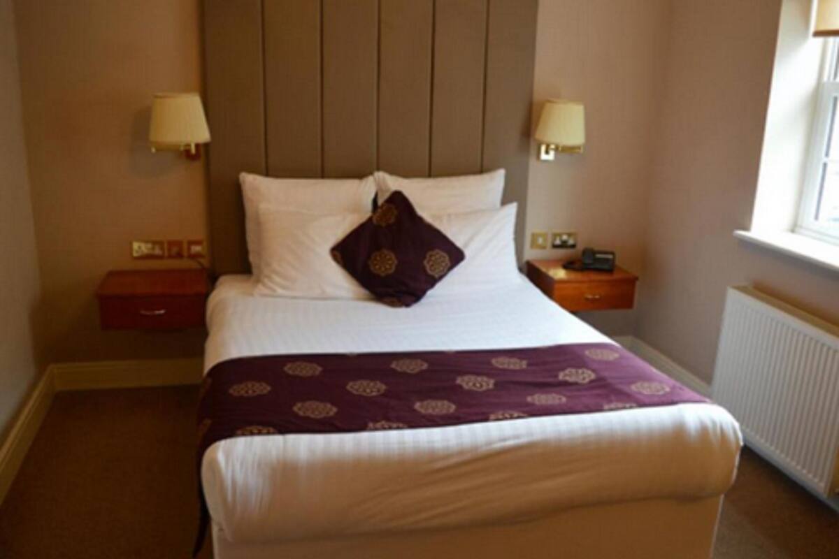 A neatly made bed with white linens and a purple patterned throw, a bedside table with a lamp, and a window with a view.