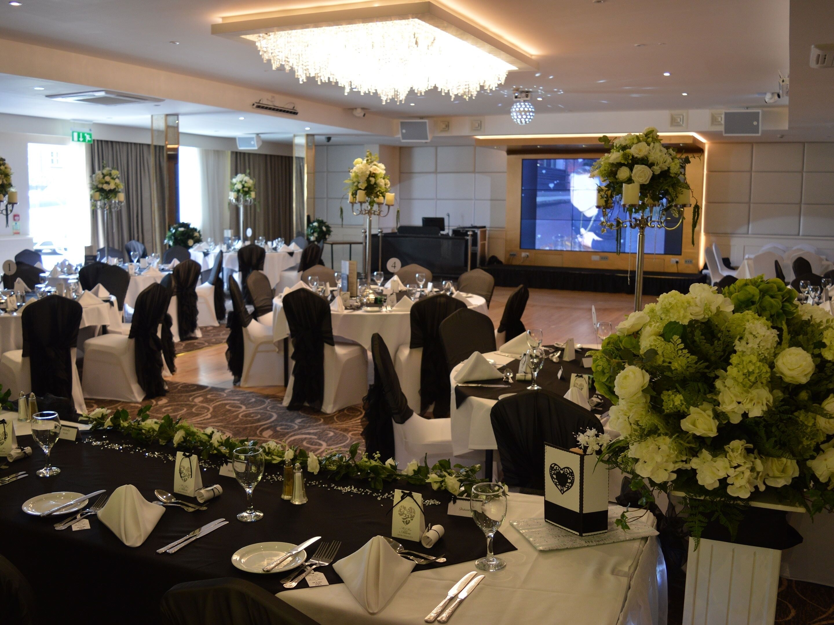 A banquet hall with round tables set for a formal event, featuring white tablecloths, black chair covers, and floral centerpieces.