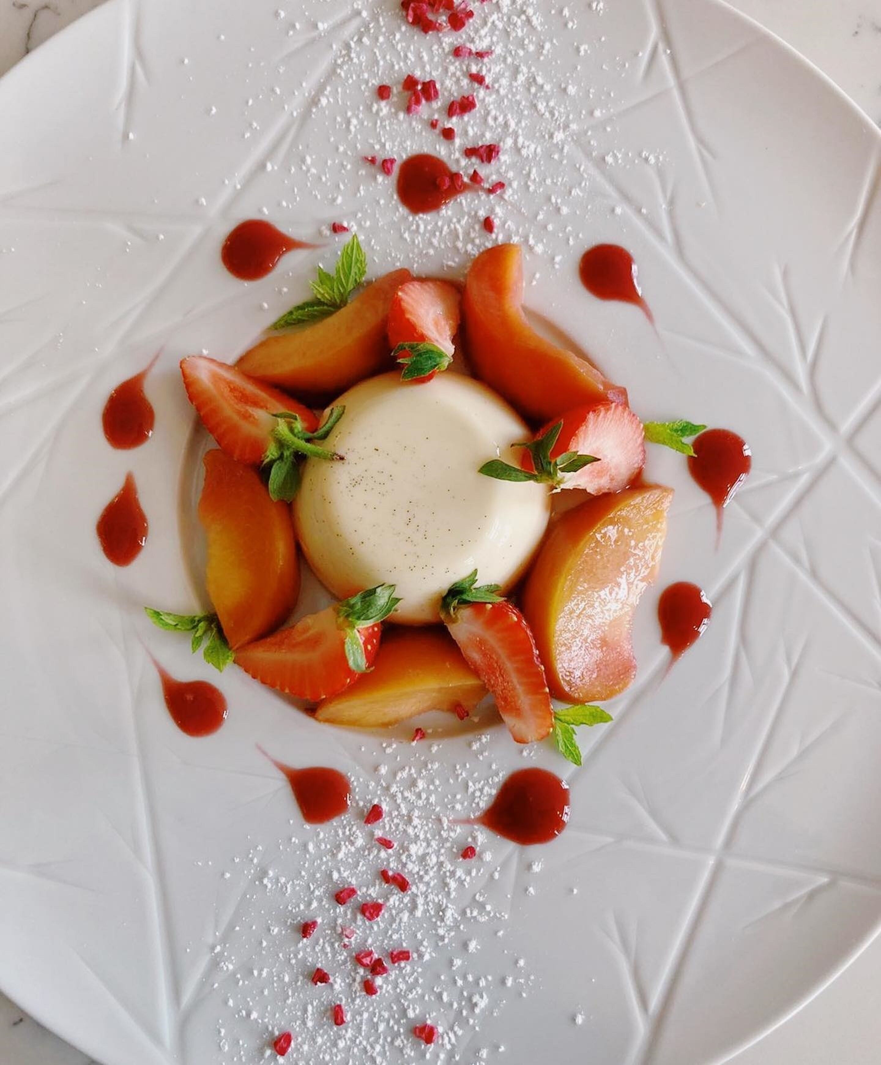 Een wit bord met een dessert bestaande uit een bolletje ijs, omringd door perzikpartjes en aardbeien, versierd met muntblaadjes en rode suikerkristallen.