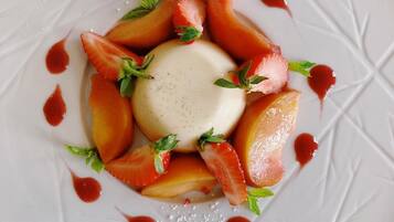 Een wit bord met een dessert bestaande uit een bolletje ijs, omringd door perzikpartjes en aardbeien, versierd met muntblaadjes en rode suikerkristallen.