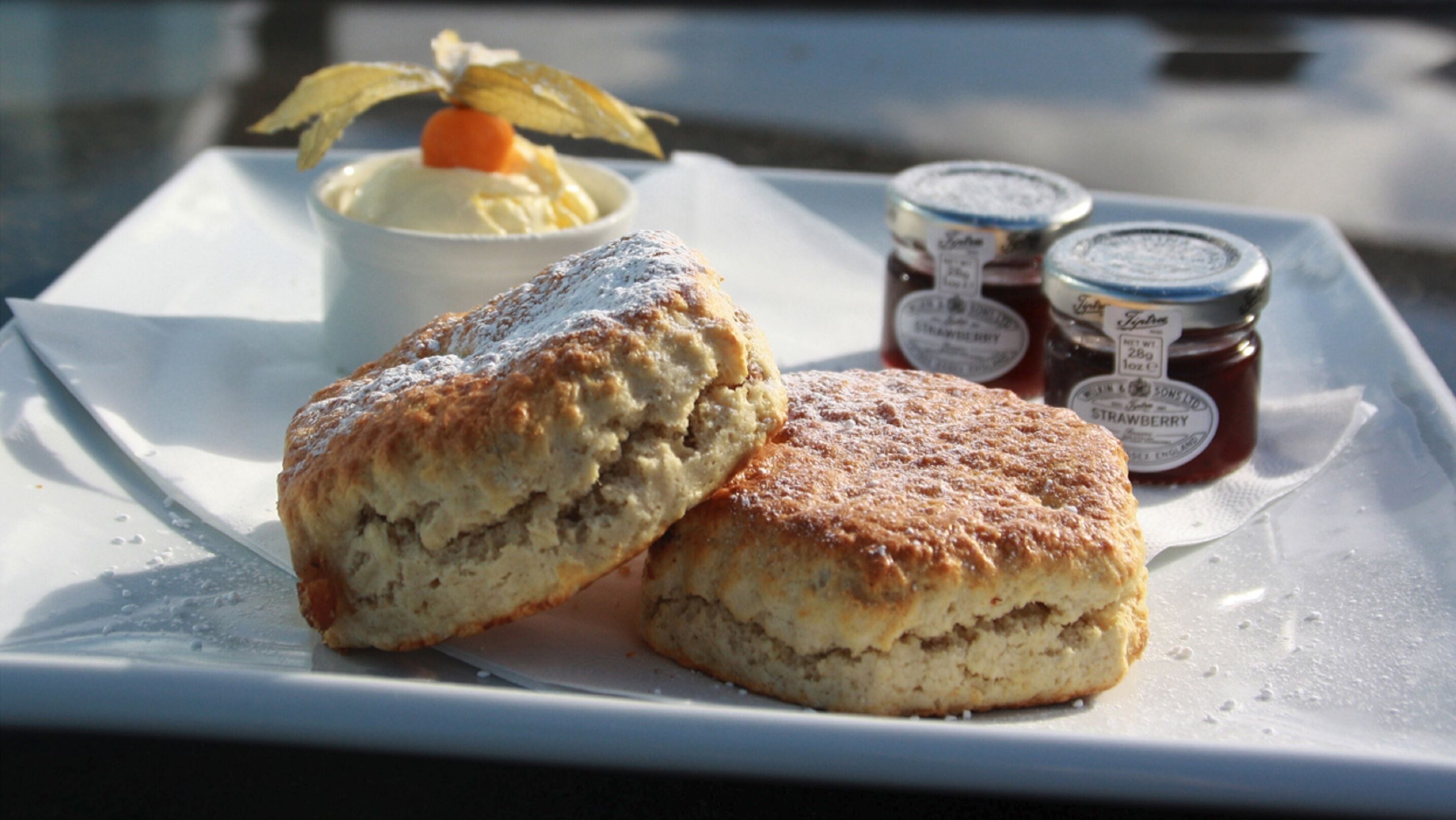 Two scones on a white plate with a small bowl of cream and two jars of jam.