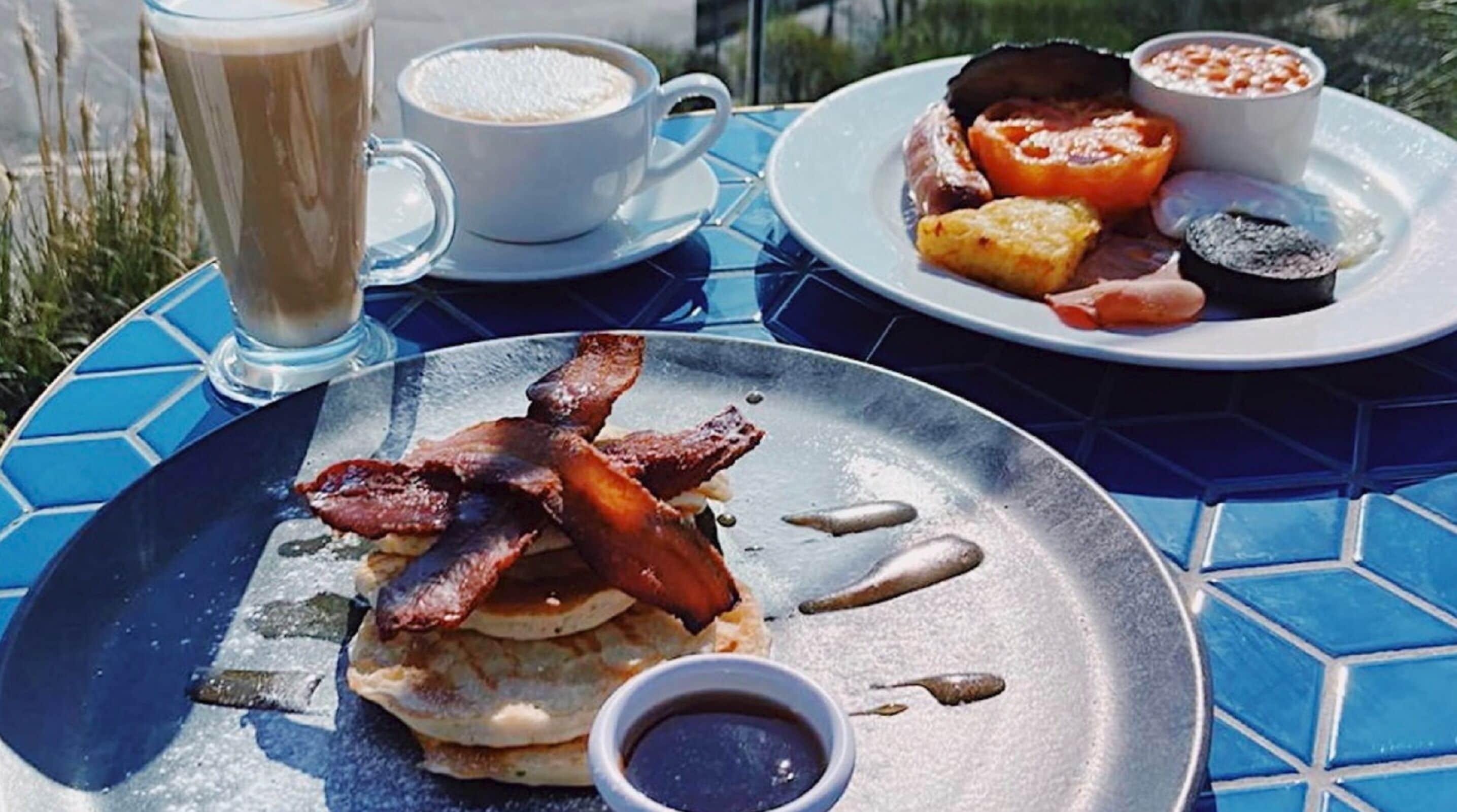 A breakfast setting with pancakes, bacon, eggs, and coffee on a blue-tiled table.