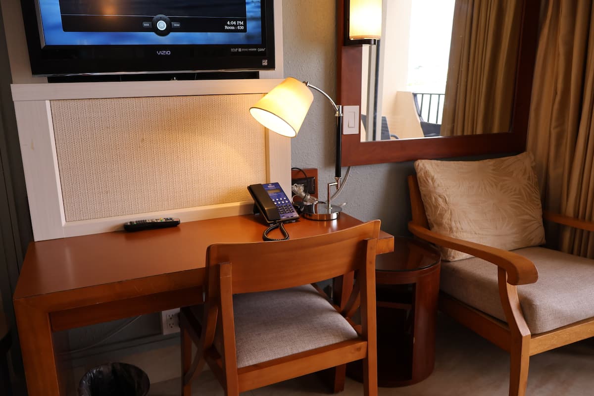 Quarto de hotel com escrivaninha de madeira, duas cadeiras, televisão, abajur e telefone.