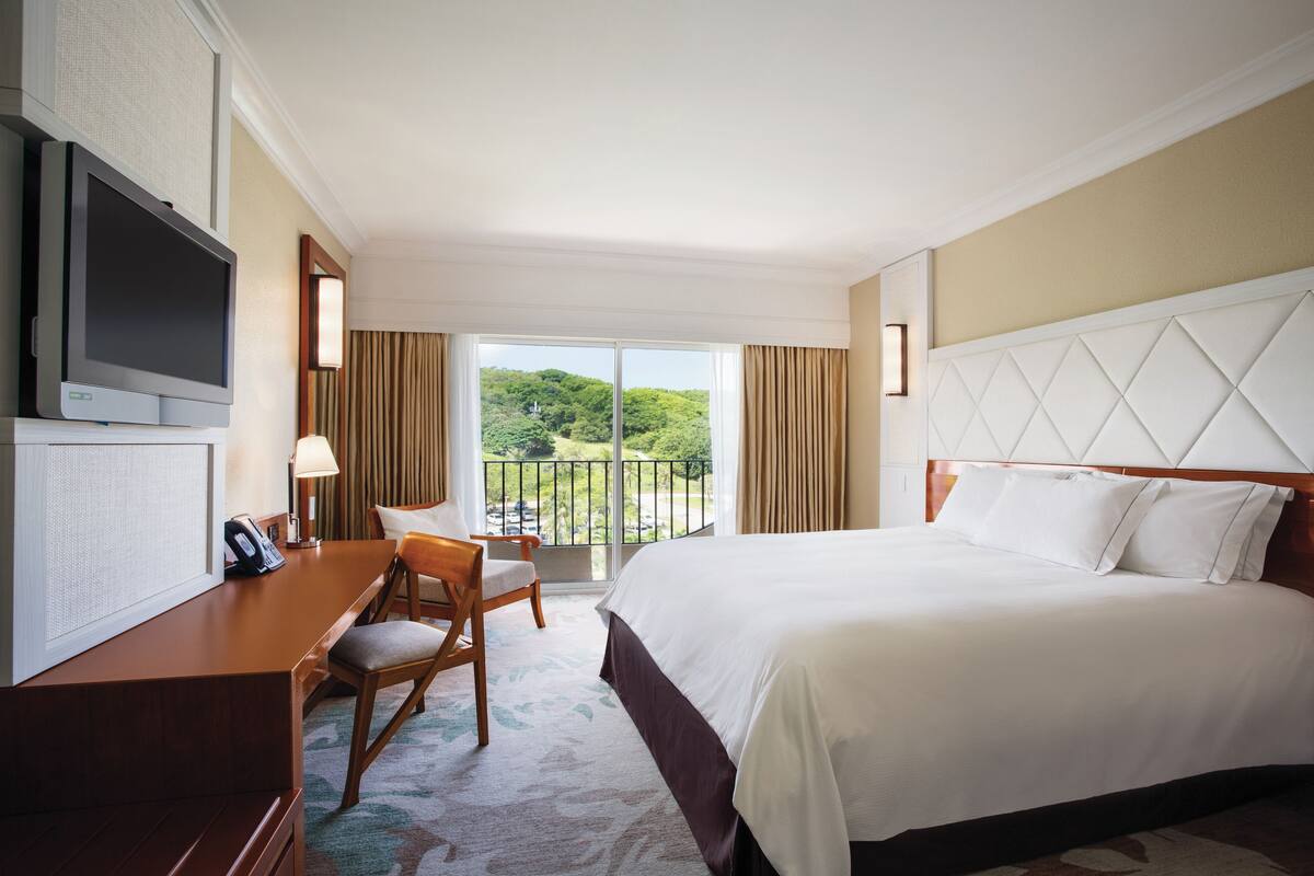 Quarto de hotel com cama, escrivaninha com cadeira, televisão e varanda com cortinas.