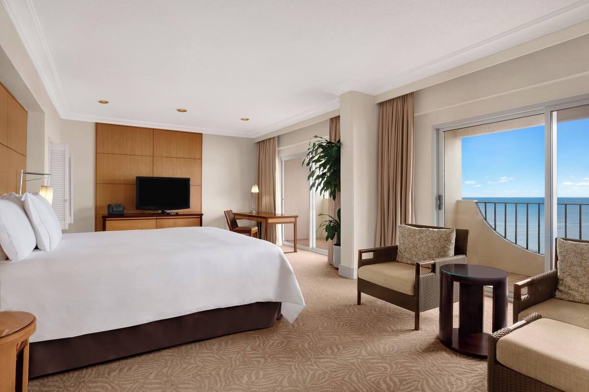 Quarto de hotel com uma cama grande, televisão, varanda com vista e área de refeições.