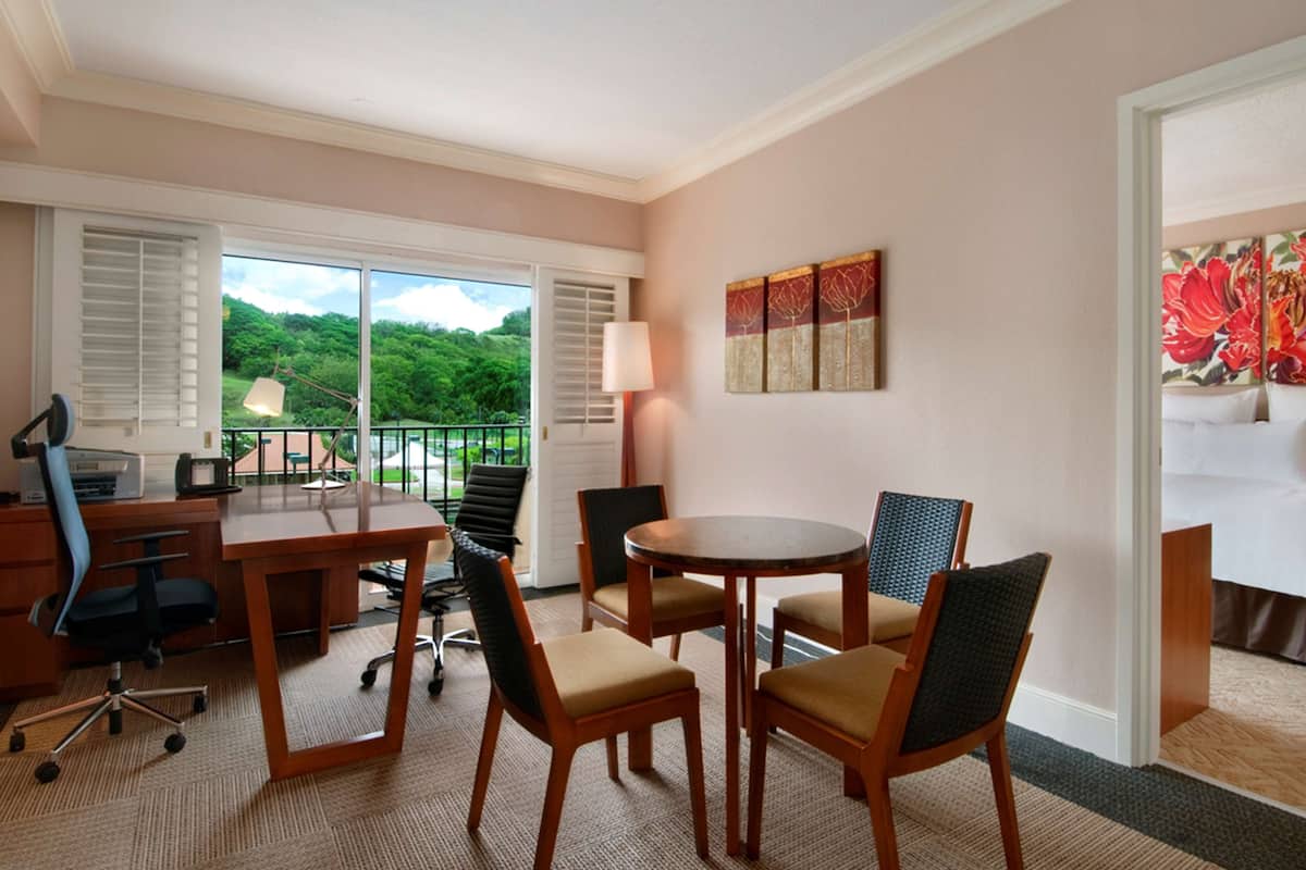 Quarto de hotel com uma janela grande, uma escrivaninha com computador, uma cadeira e uma mesa redonda com cadeiras.