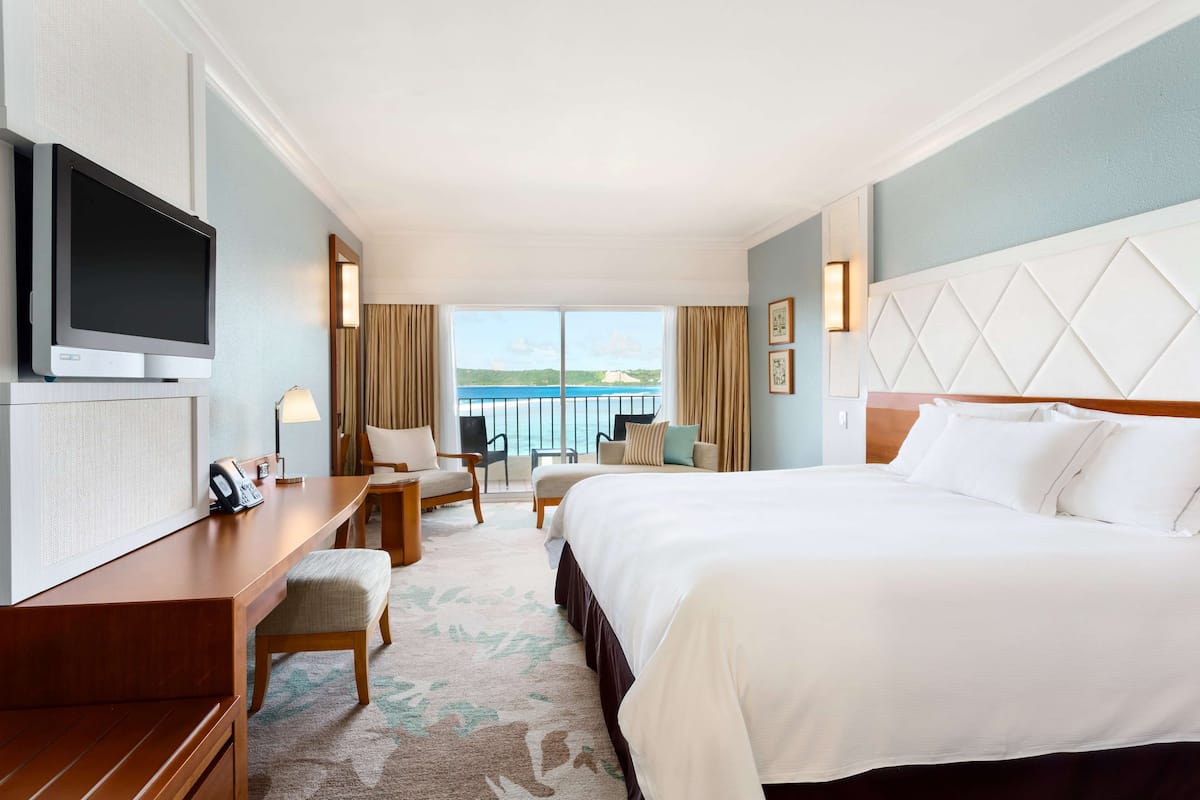 Quarto de hotel com uma cama grande, uma escrivaninha com televisão de tela plana, uma cadeira, uma mesa pequena e vista para o mar através de amplas janelas.