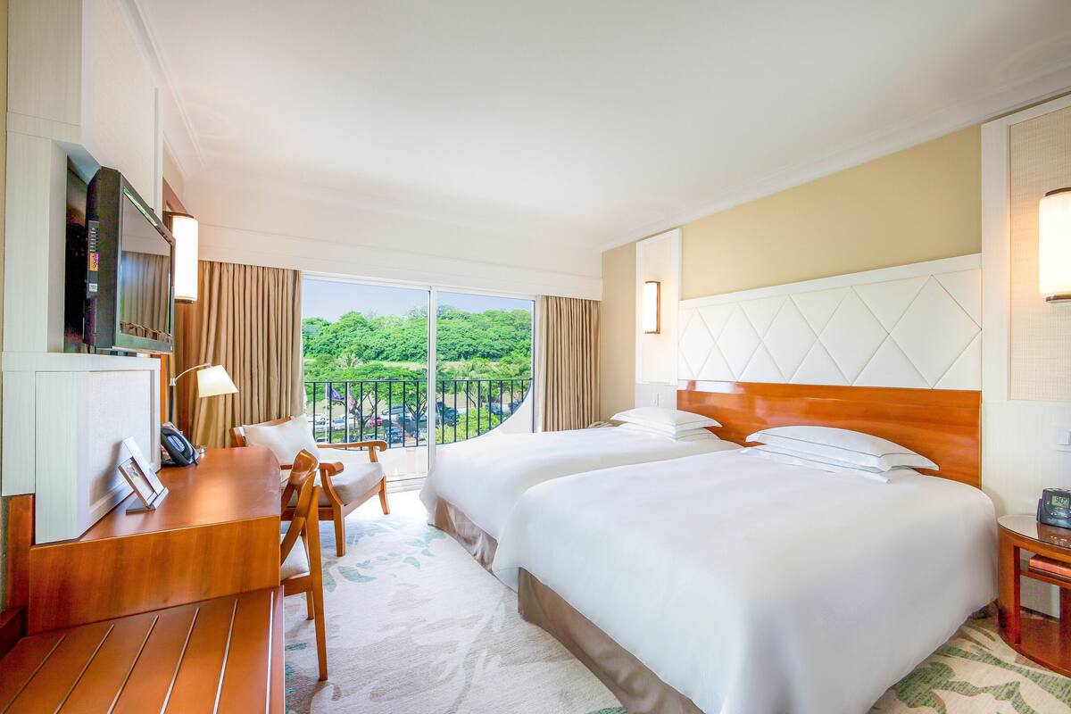 Quarto de hotel com duas camas, uma escrivaninha, uma cadeira, televisão e varanda com vista para o verde.