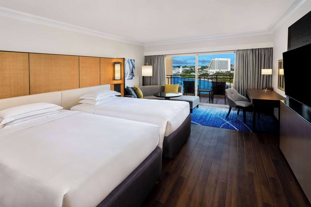 Quarto de hotel com duas camas, uma escrivaninha, televisão e varanda com vista para a cidade.