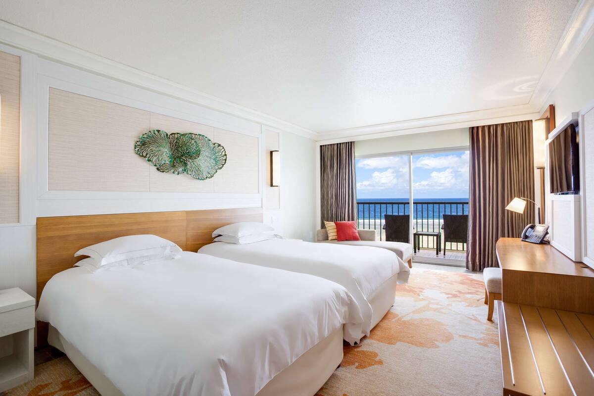 Quarto de hotel com duas camas, uma escrivaninha, uma cadeira e varanda com vista para o mar.