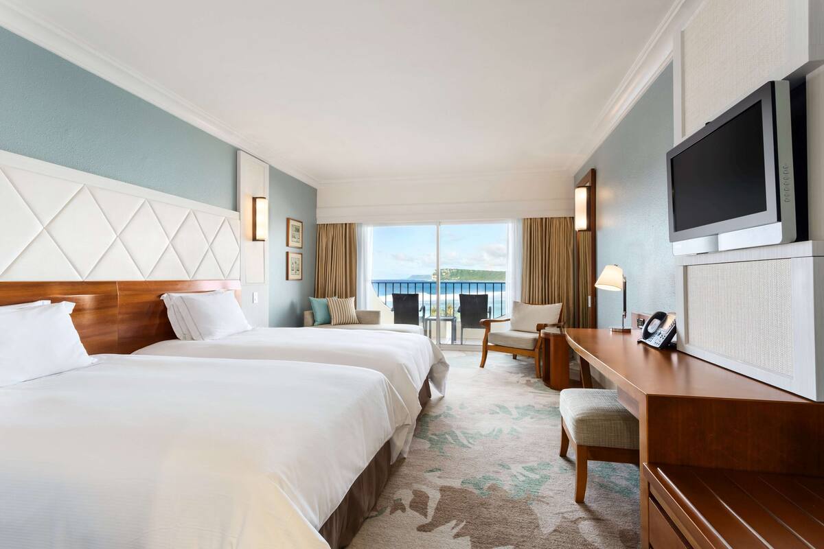 Quarto de hotel com duas camas, uma escrivaninha, uma cadeira, televisão e vista para o exterior.