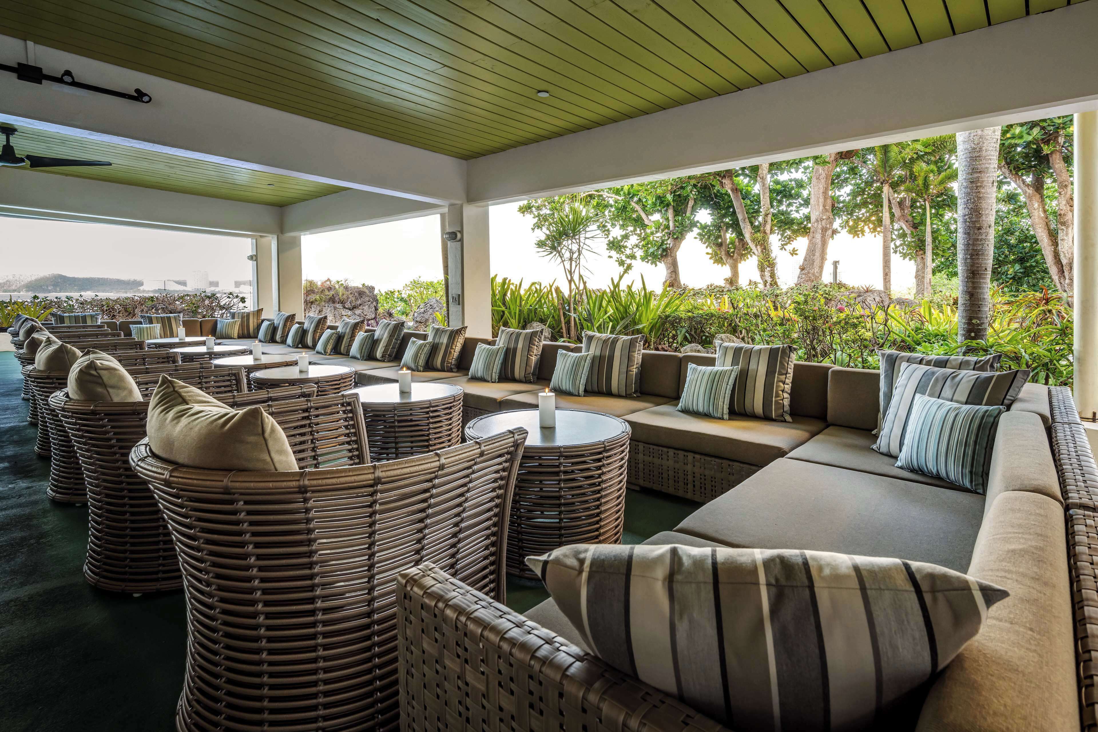 A covered outdoor seating area with wicker furniture and cushions, overlooking a landscaped garden and distant view.