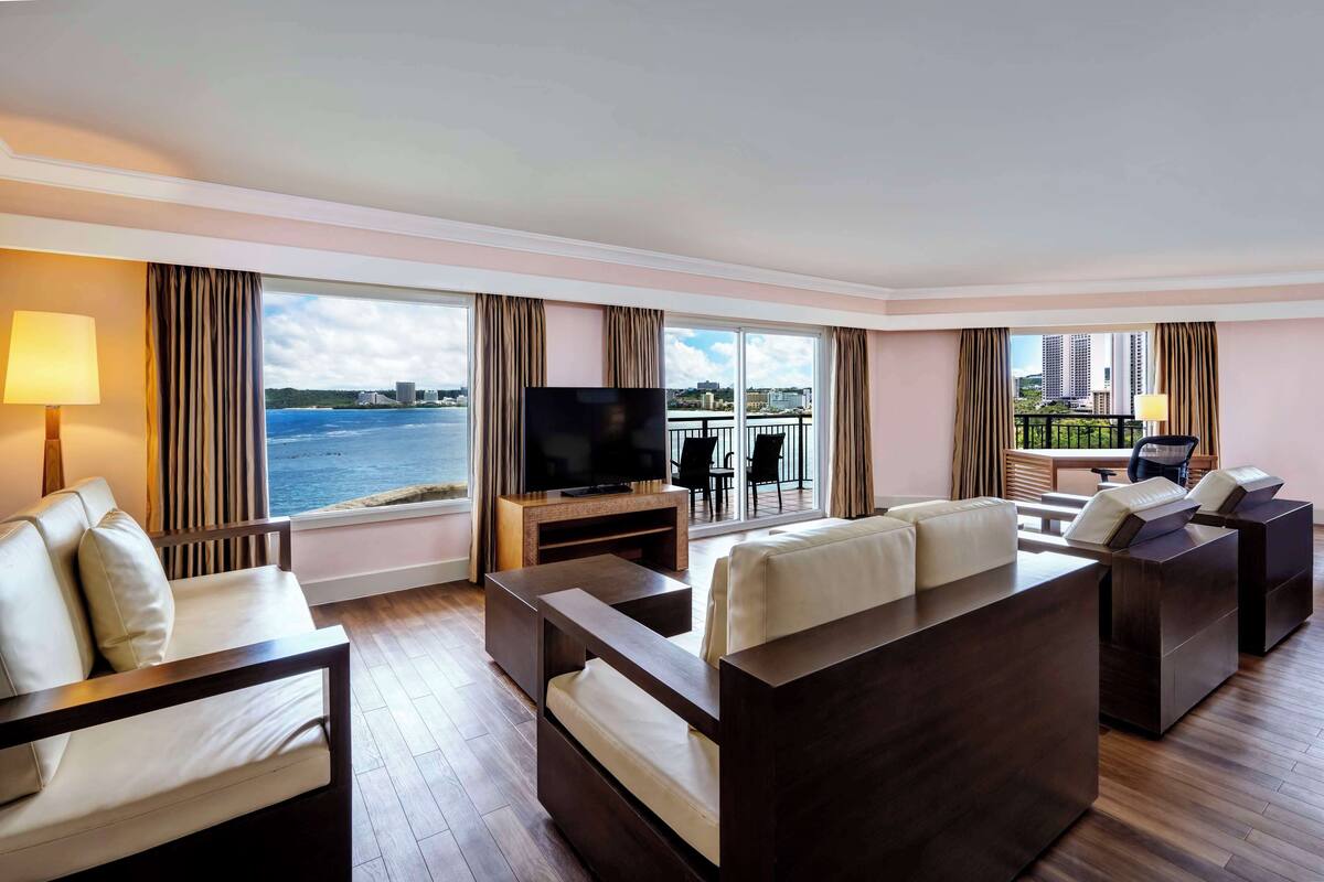 Quarto de hotel moderno com uma grande janela que oferece vista para o mar e para a paisagem urbana. Há uma televisão de tela plana em um suporte de madeira, um sofá e uma mesa pequena.