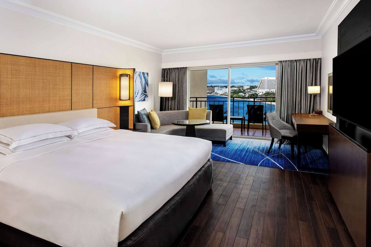 Quarto de hotel com uma cama grande, um sofá, uma escrivaninha e uma varanda com vista para a cidade.