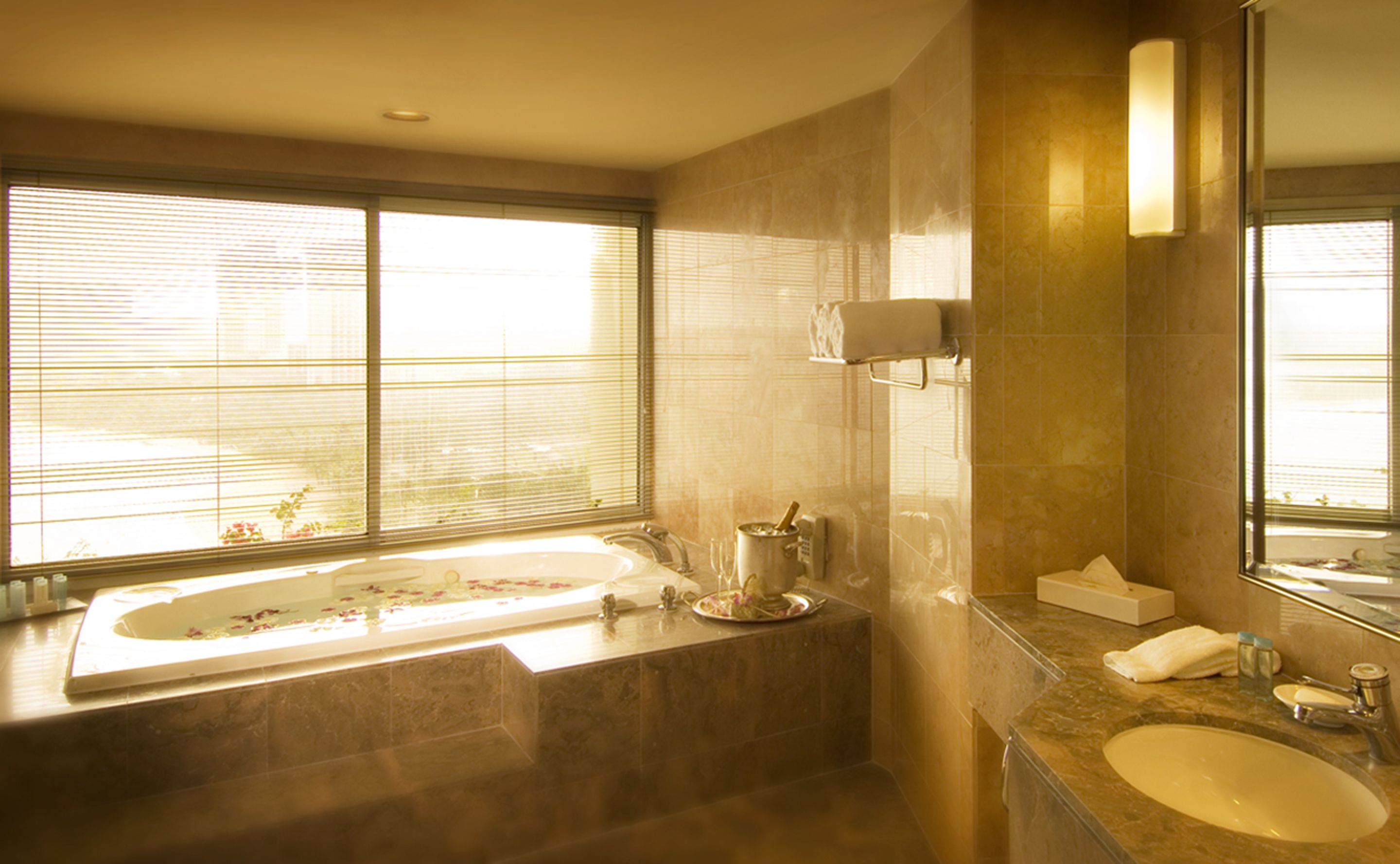 A bathroom with a large bathtub, a sink, and a mirror. There are windows with blinds, and a towel rack is visible.
