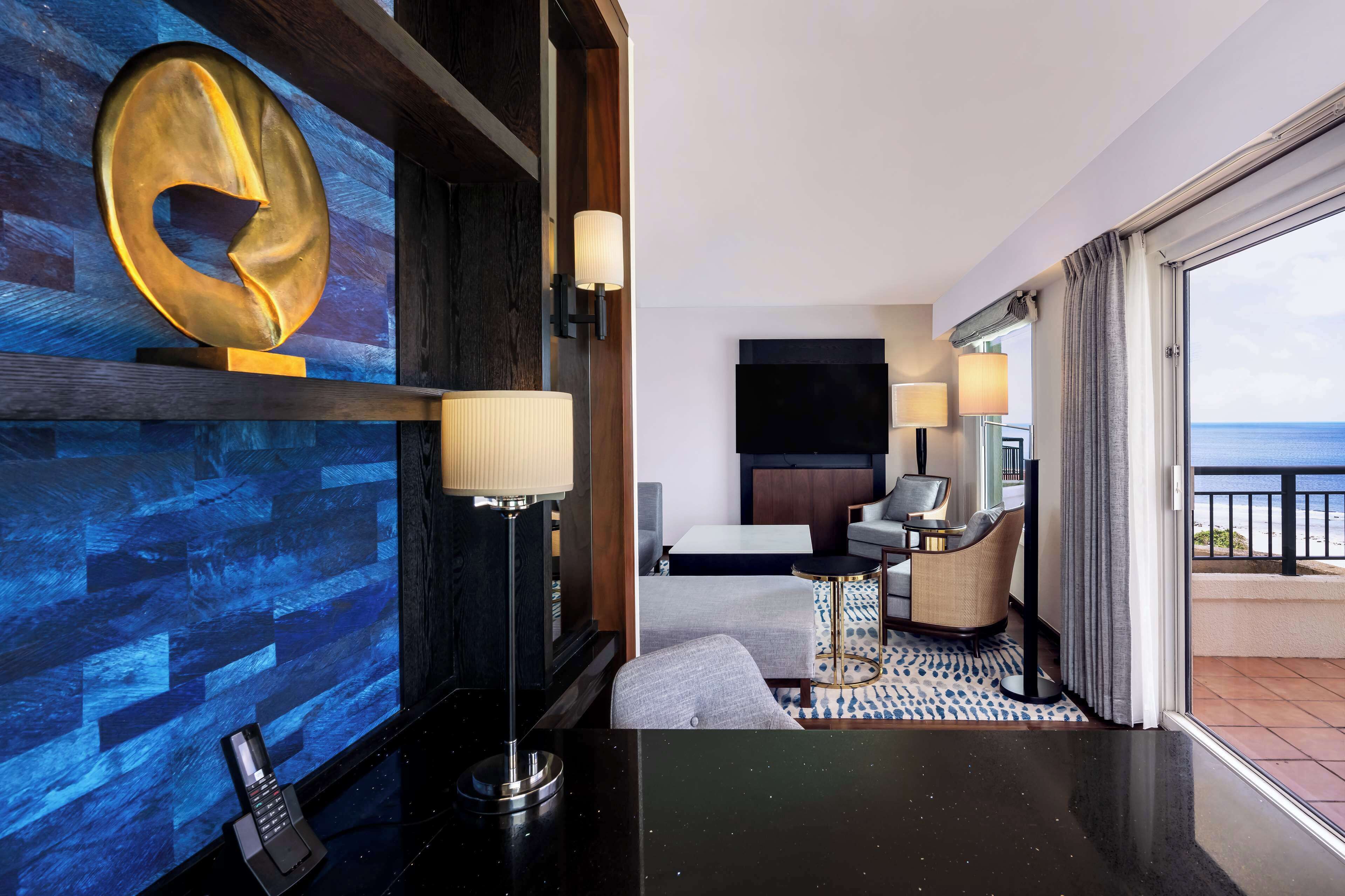 A modern hotel room with a large window, a flat-screen TV, a sofa, a desk, and a unique wall art piece.