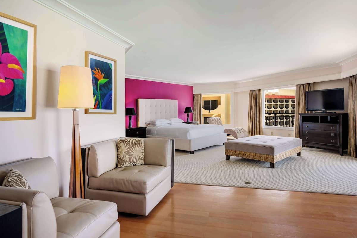Quarto de hotel com cama, sofá, televisão e área de refeições com mesa e cadeiras.