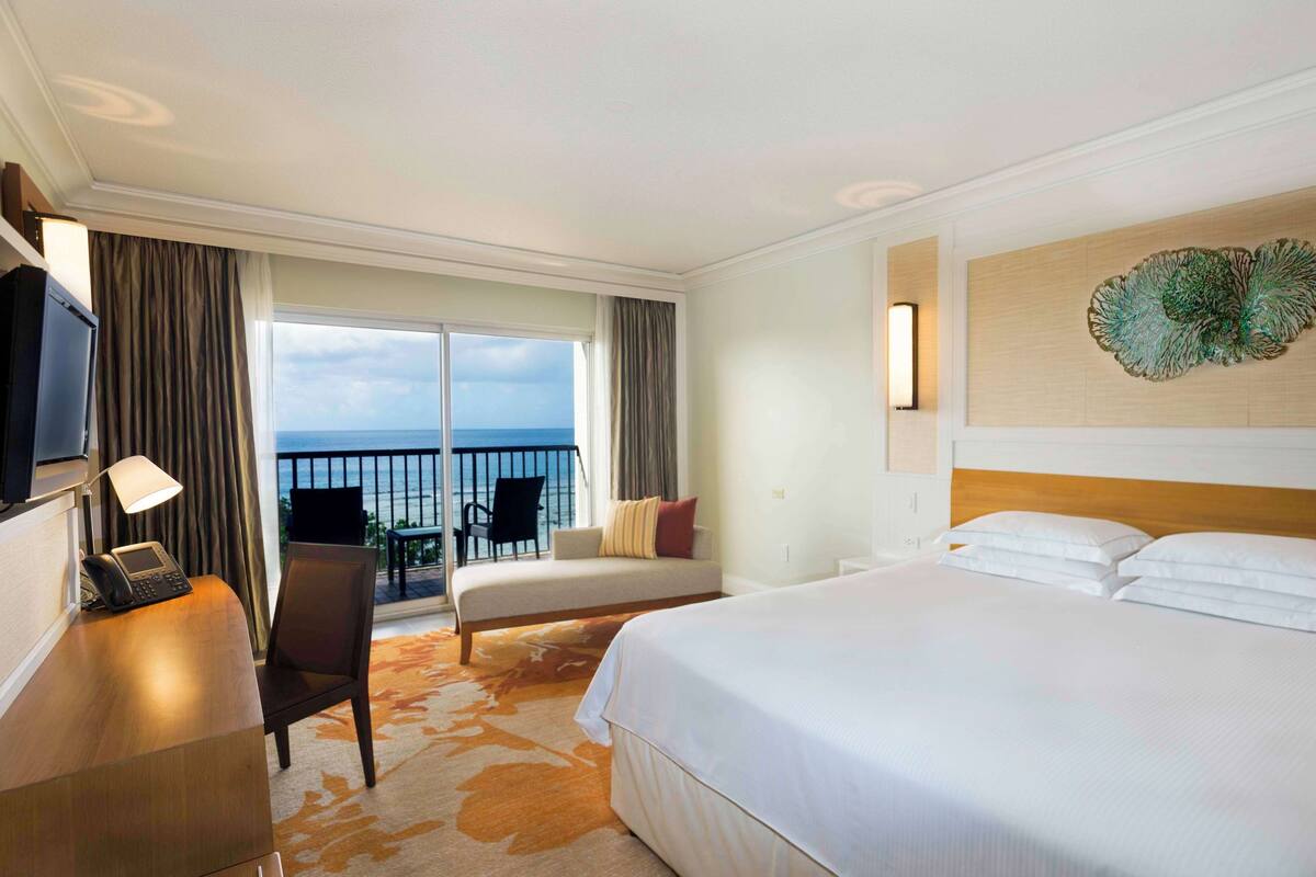 Quarto de hotel com uma cama grande, uma escrivaninha com telefone, uma cadeira, uma varanda e vista para o mar, e um objeto decorativo na parede.