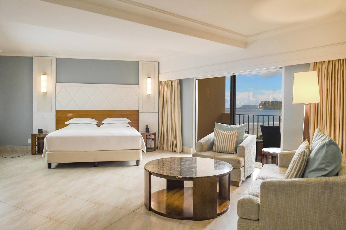 Quarto de hotel com cama grande, mesa de centro redonda, sofá e vista para o mar.