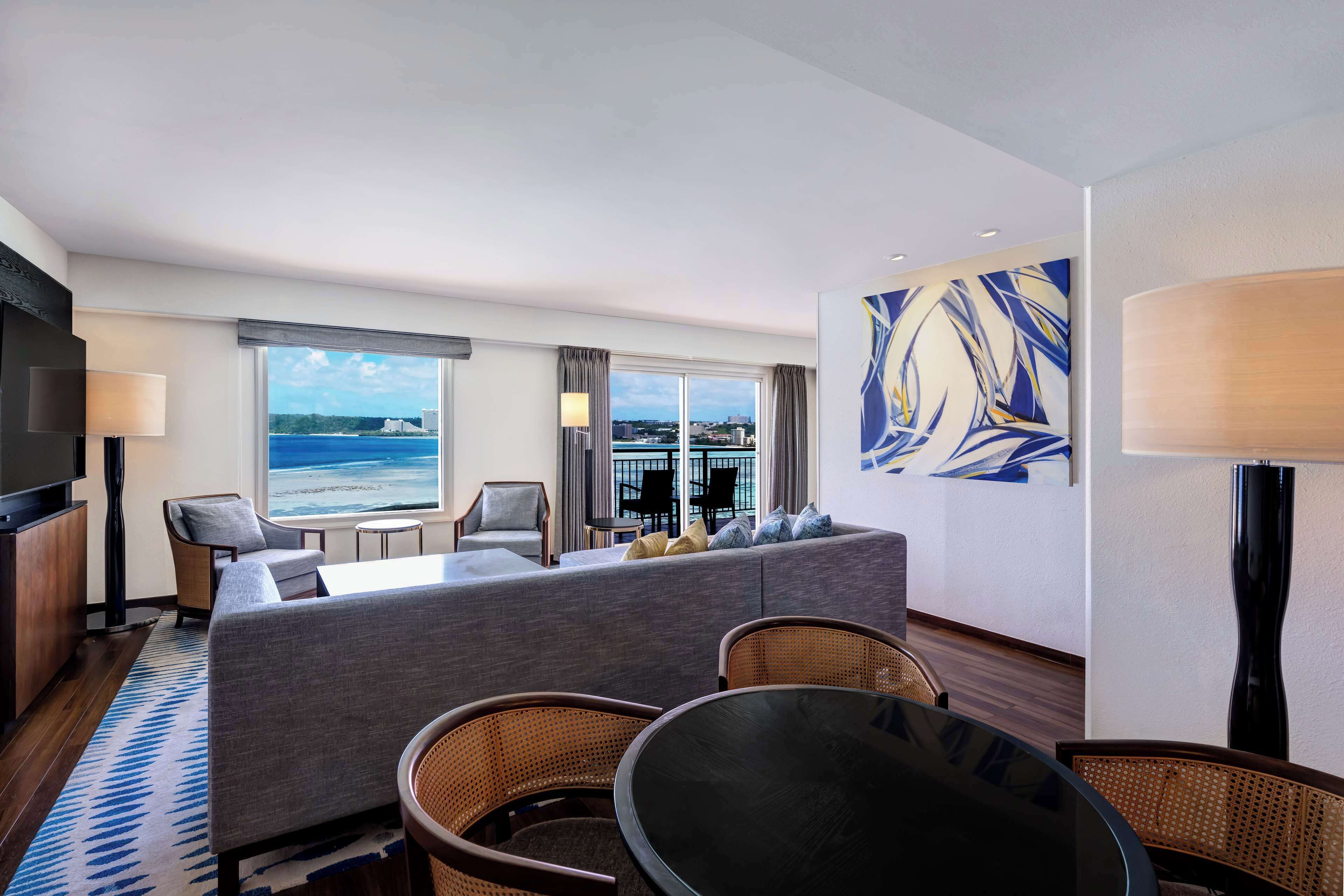 A hotel room with a large window offering a view of the ocean, a sofa, a dining table, and a flat-screen TV.