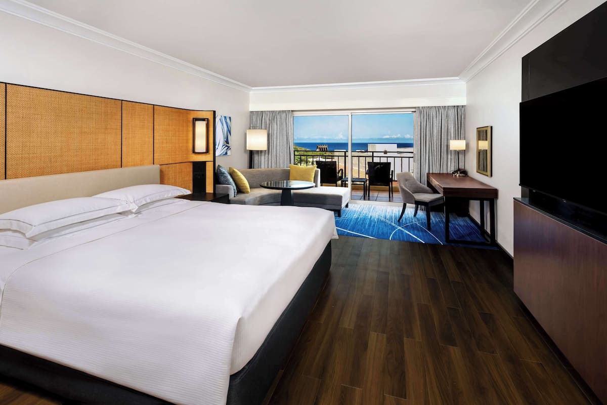 Quarto de hotel com uma cama grande, uma escrivaninha, uma cadeira, uma televisão e varanda com vista.