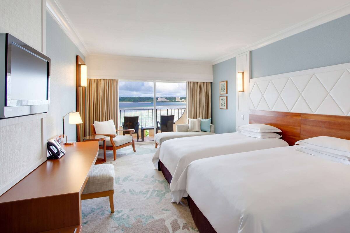 Quarto de hotel com duas camas, uma escrivaninha com telefone, uma cadeira, televisão e varanda vista para o mar.