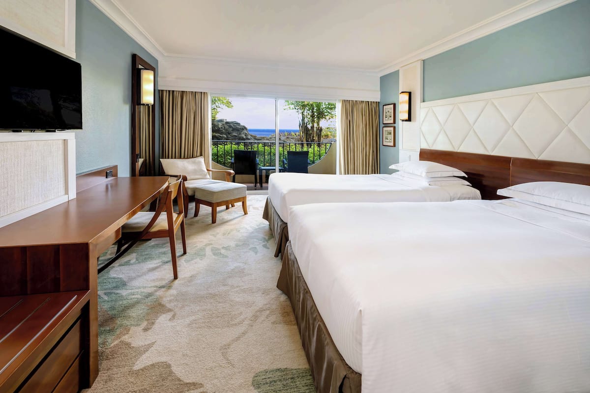 Quarto de hotel com duas camas, uma mesa, televisão e varanda com vista para o mar.