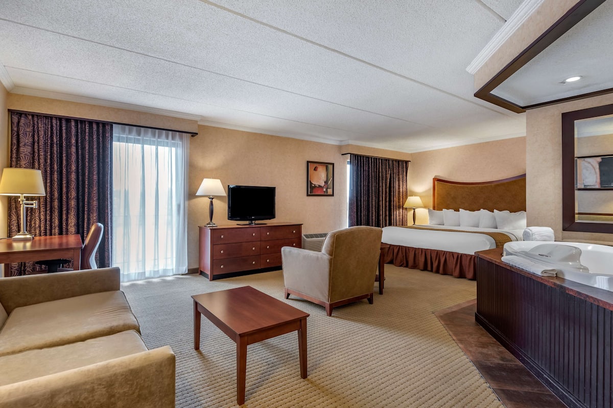 A hotel room with a bed, a desk with a lamp, a chair, a sofa, a TV, and a bathroom with a bathtub.