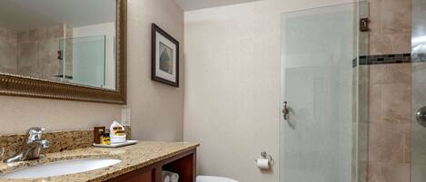 A bathroom with a granite countertop sink, a white toilet, a framed mirror, and a glass shower enclosure.