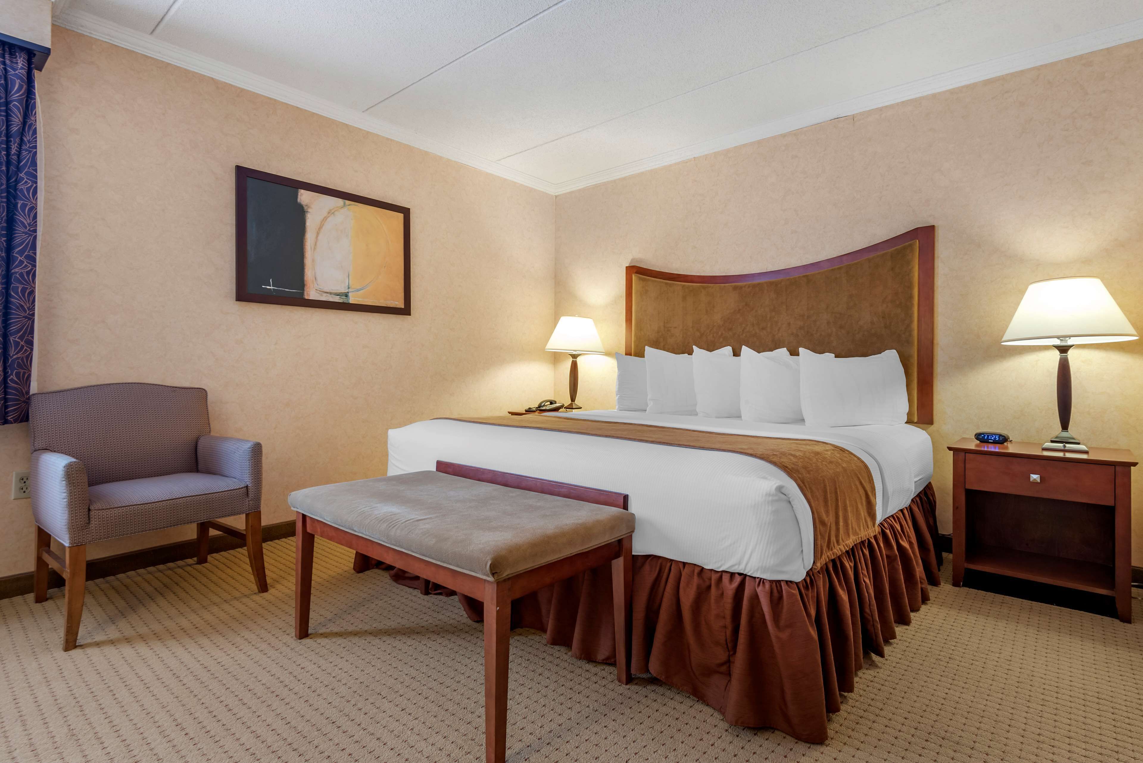 A hotel room with a large bed, a chair, a bench, two bedside lamps, and a painting on the wall.