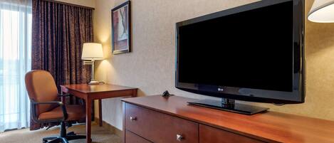 Quarto de hotel com televisão de tela plana, escrivaninha de madeira, cadeira, abajur e quadro na parede.