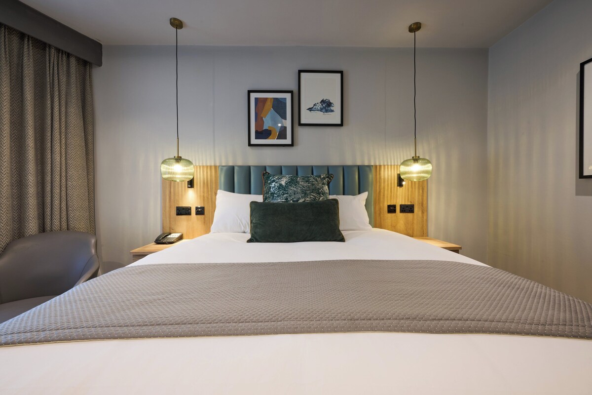 A hotel room with a large bed, two bedside lamps, a headboard, a pillow, a bedside table with a phone, and a chair.