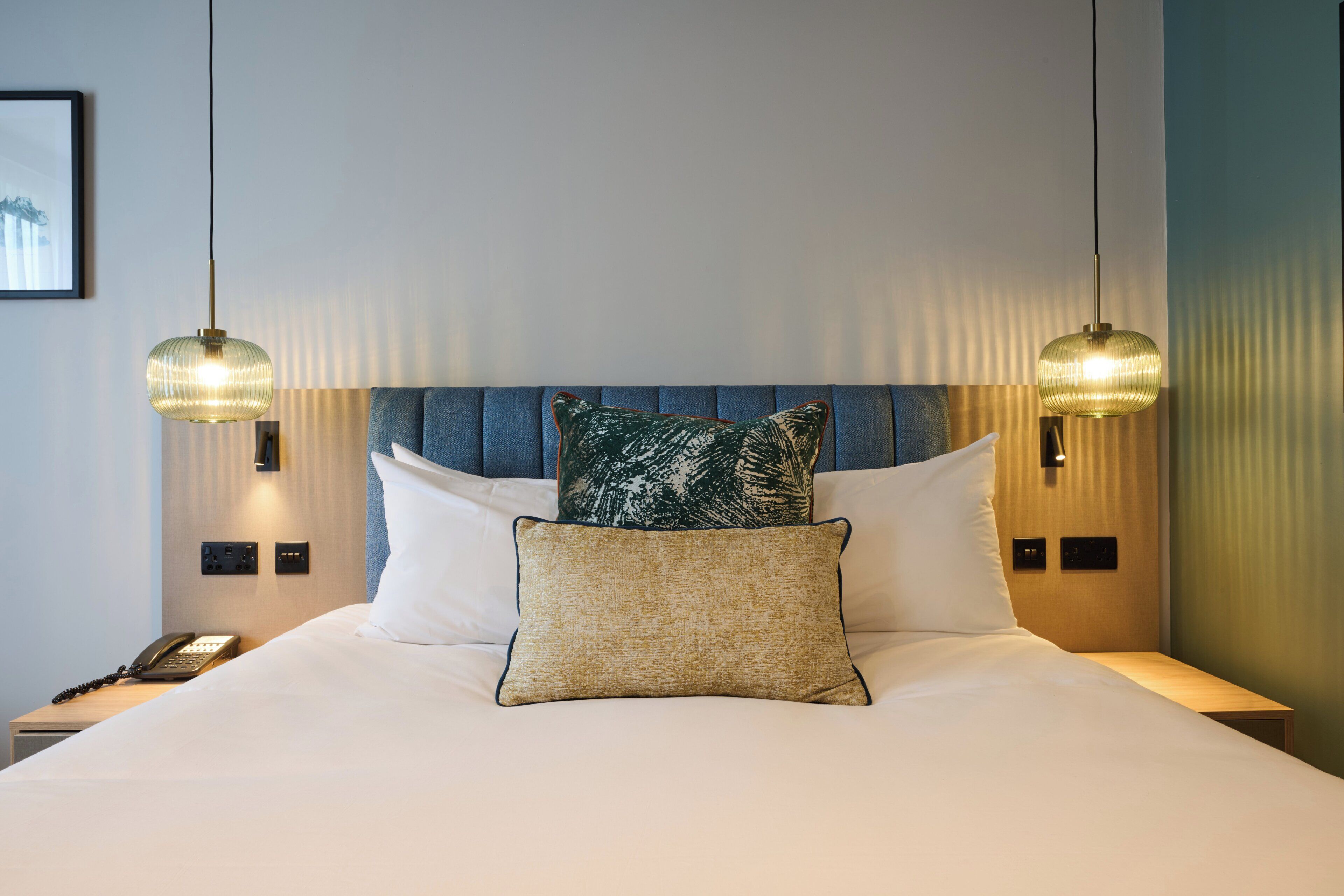 A hotel room with a bed, two bedside lamps, a telephone, and a wall-mounted light switch.
