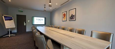 A conference room with a long table, chairs, a projector screen, and framed artwork on the wall.