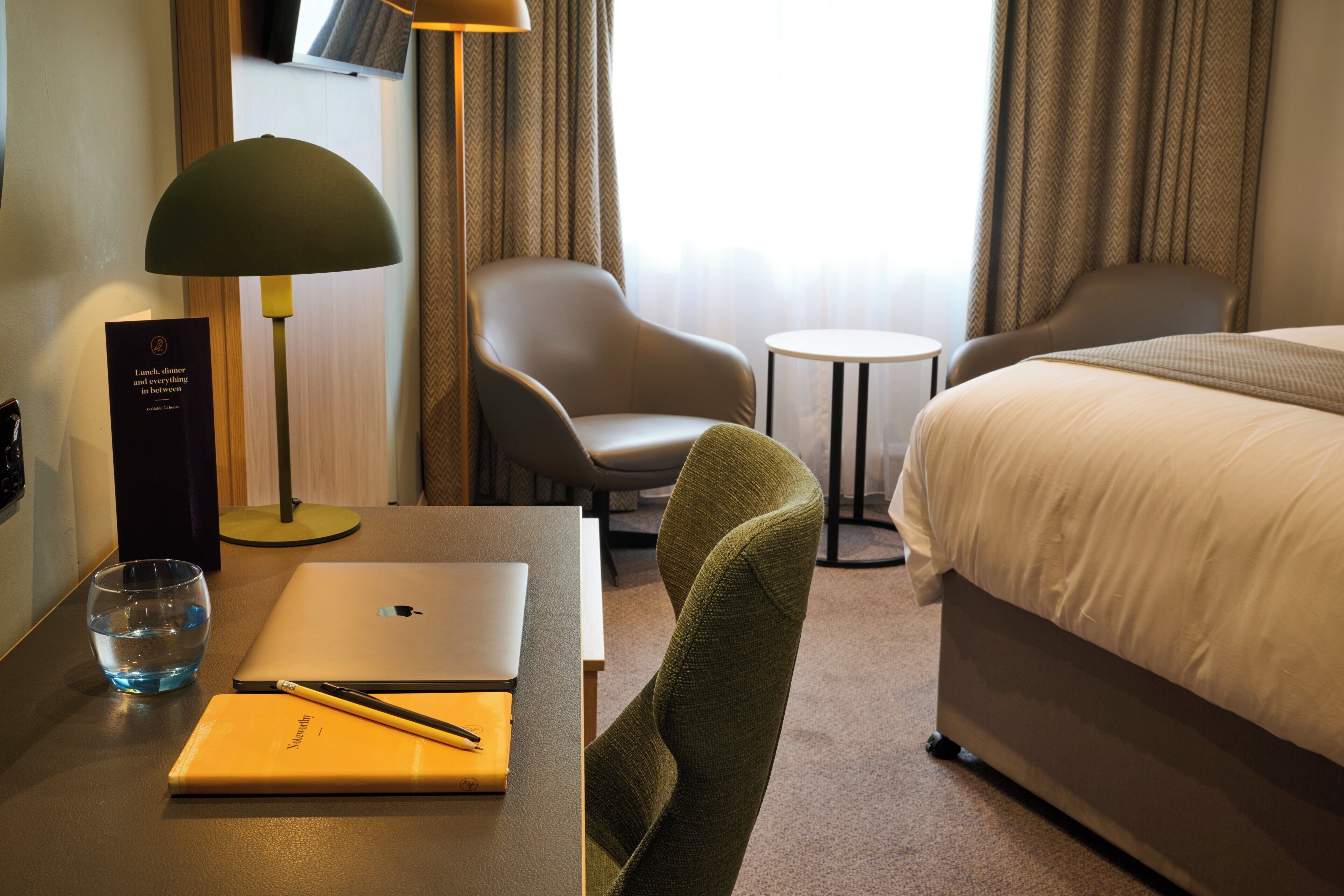 Et hotelværelse med seng, skrivebord med computer, stol, lampe og et glas vand.