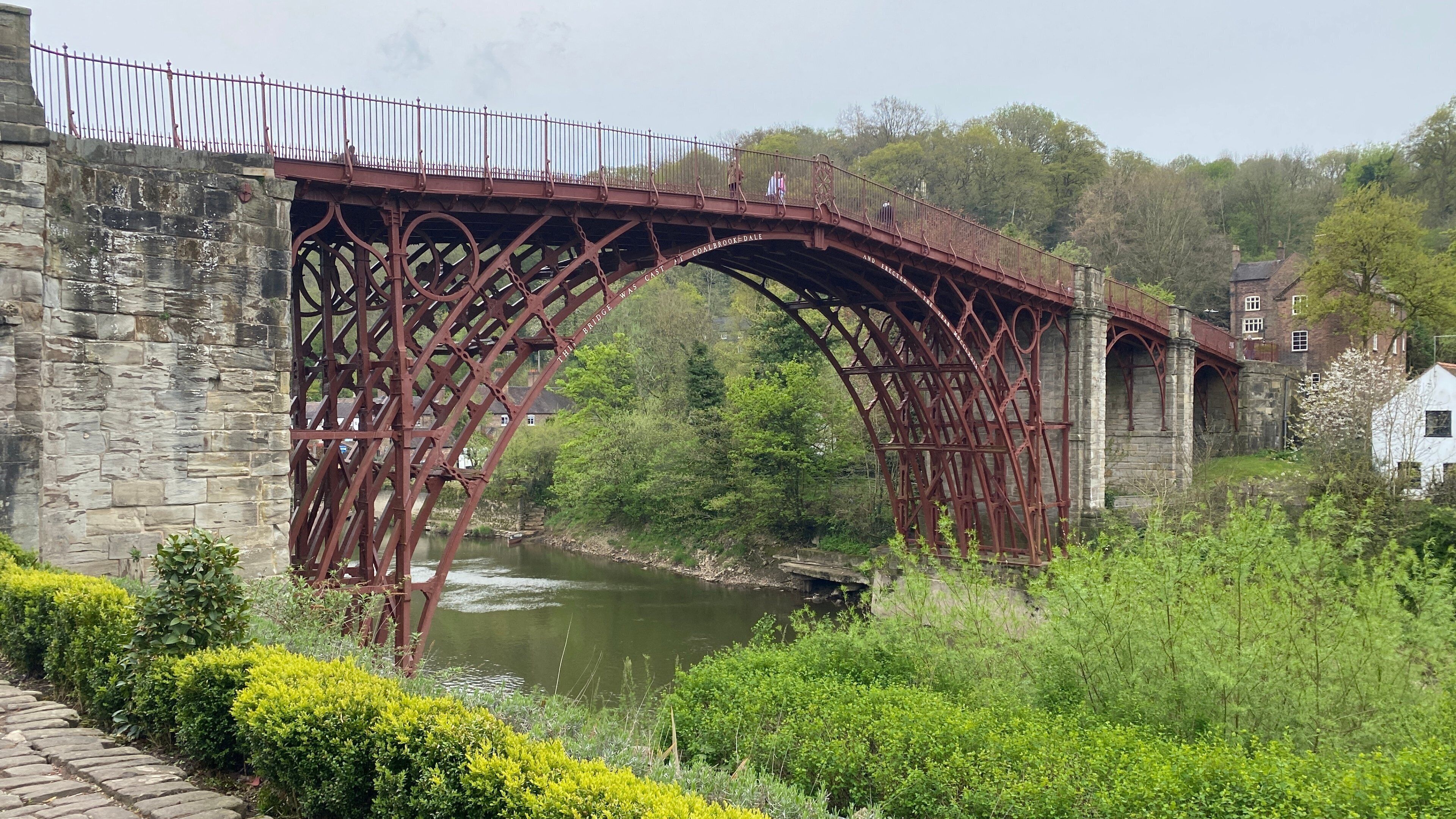 A historic iron bridge with stone walls on either side, a river below, and a building with a red roof in the background.