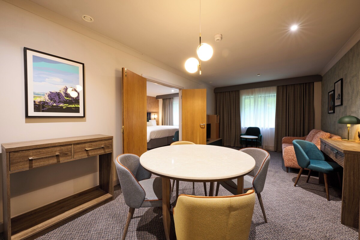 A hotel room with a bed, a desk, a round table with chairs, a sofa, and a picture on the wall.