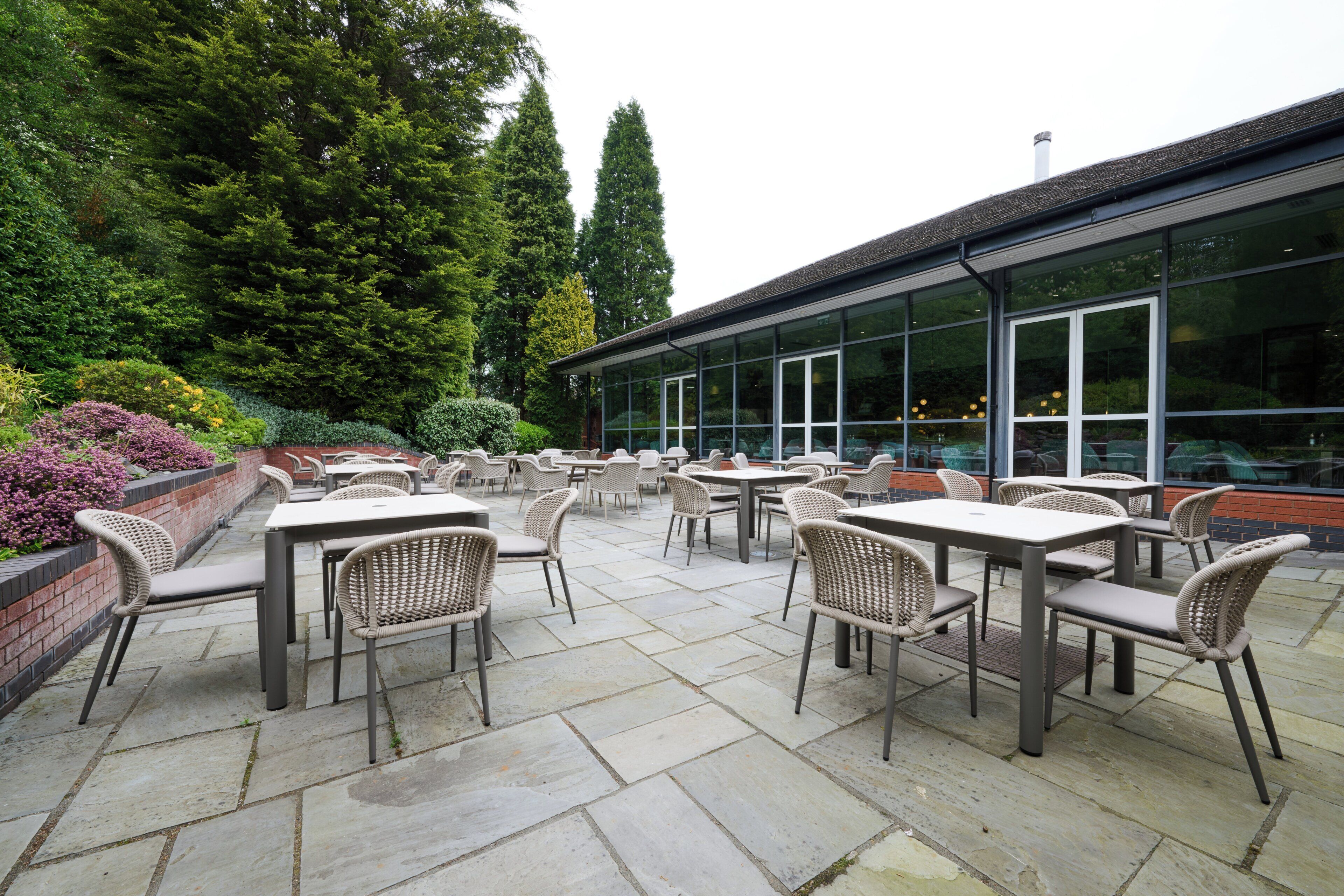 Un espace extérieur aménagé en terrasse, avec des tables et des chaises, entouré de verdure et bordé d’un bâtiment doté de grandes fenêtres.