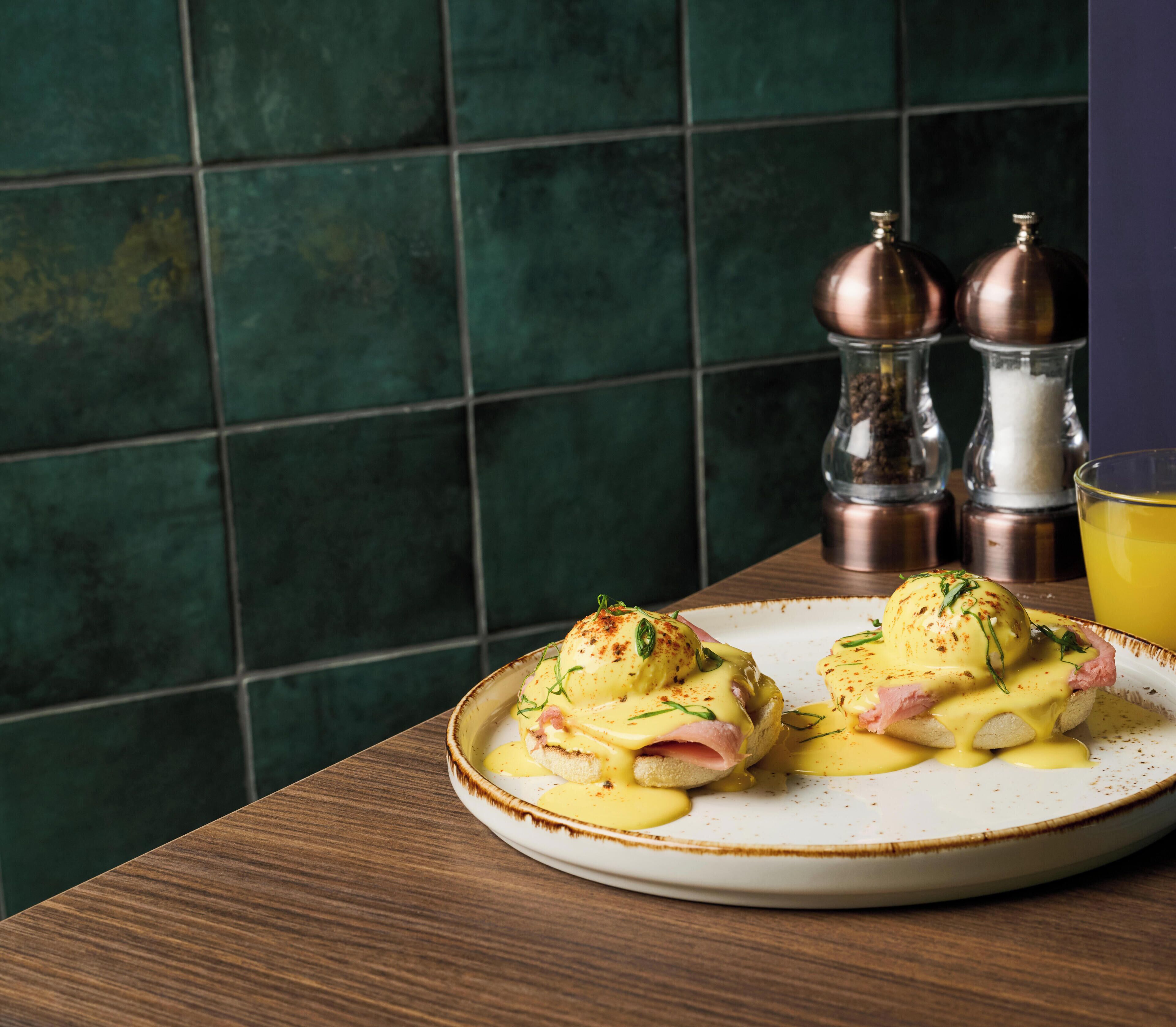 Un plato de huevos benedictinos sobre un plato de cerámica, con una mesa de madera y una pared de azulejos verdes al fondo.