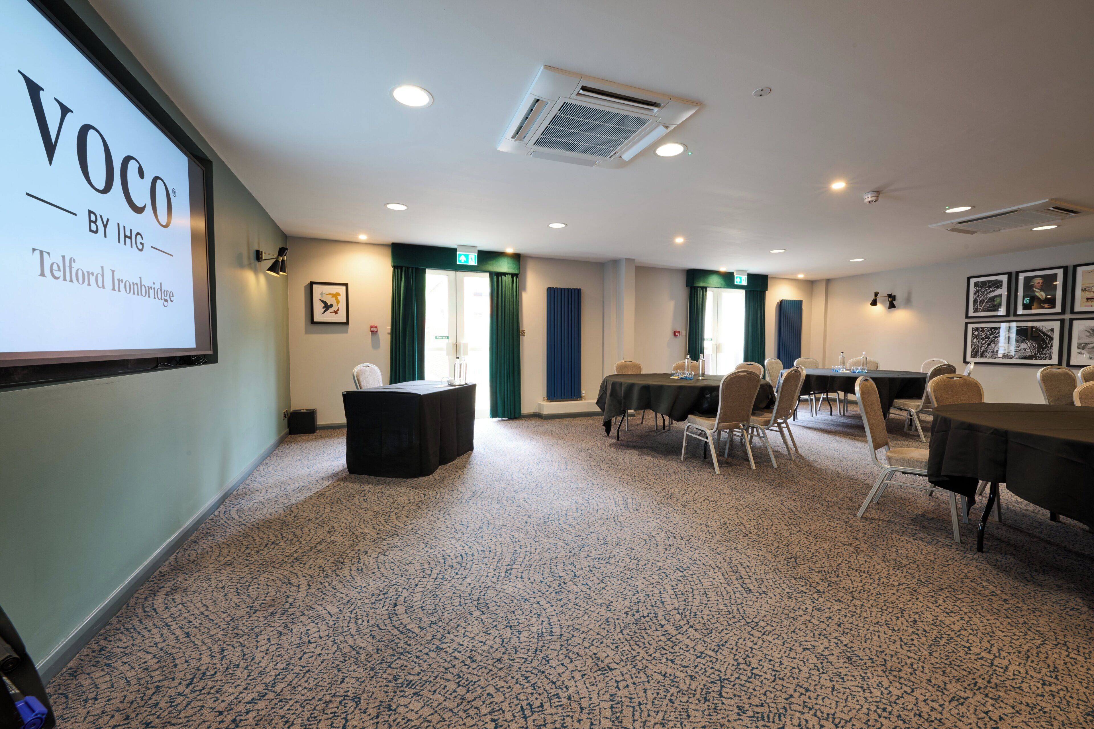 A conference room with a projector screen displaying 'Voco by IHG Telford Ironbridge,' a table with chairs, and a carpeted floor.