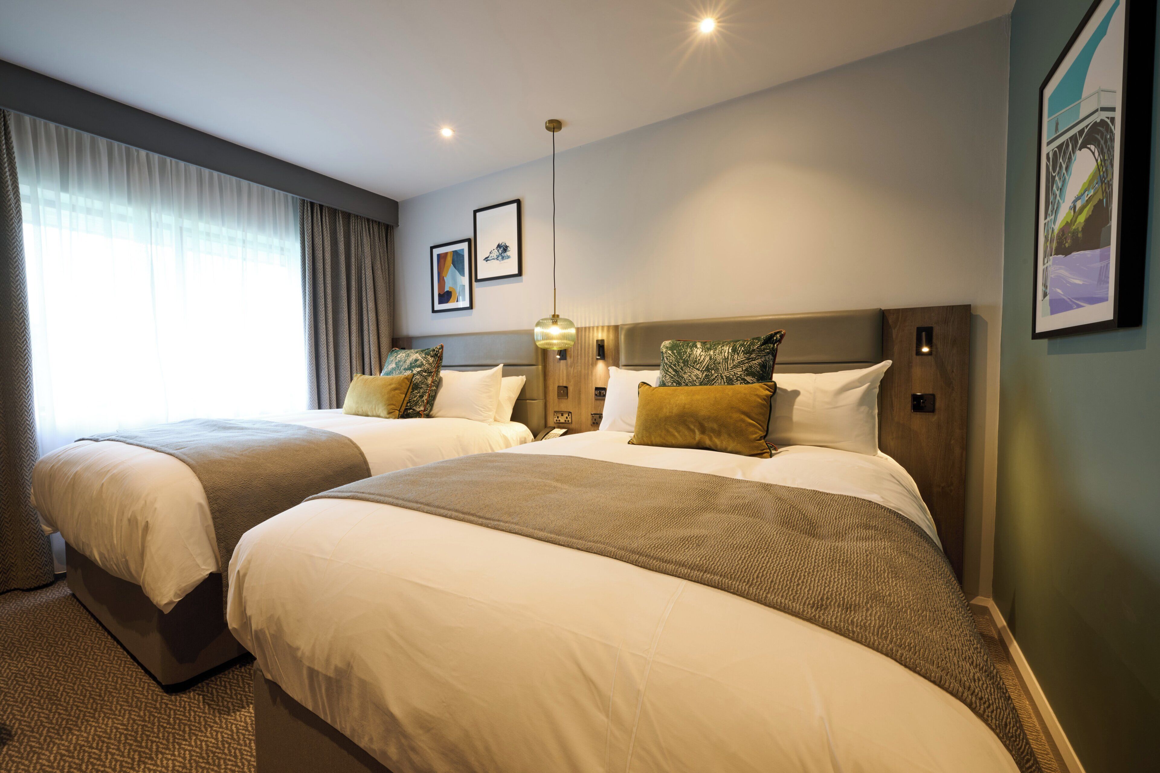 Habitación de hotel con dos camas, un ventanal con cortinas, una lámpara de pared y cuadros enmarcados.