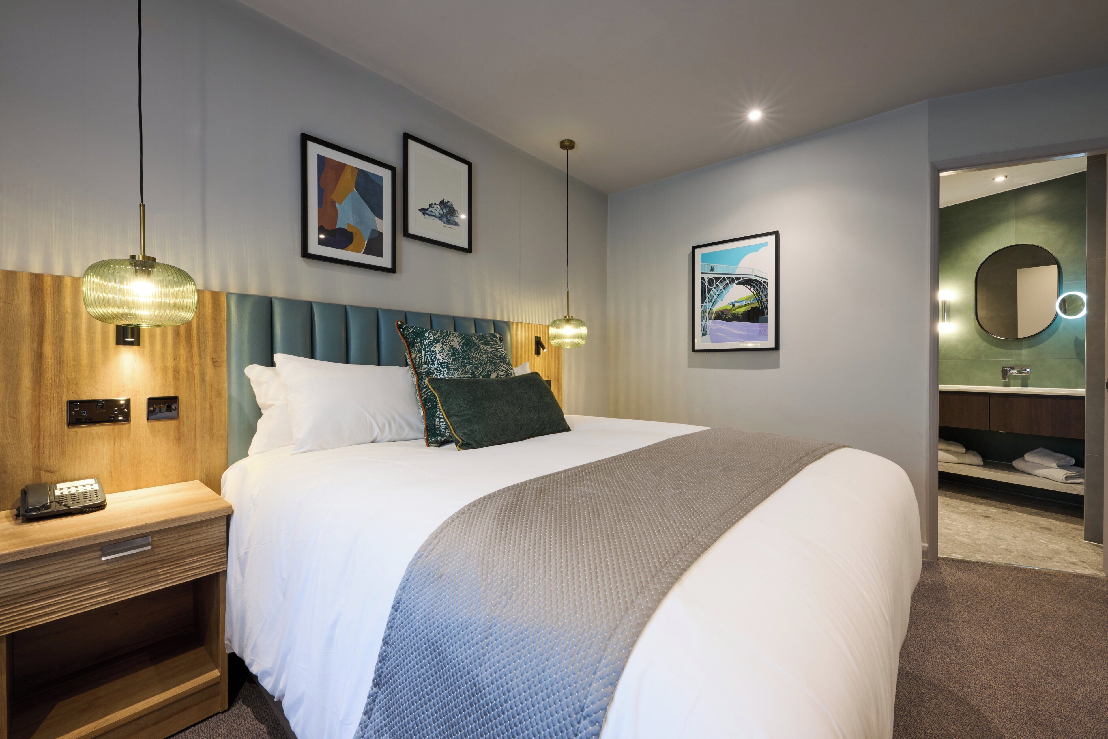 Une chambre d’hôtel avec un lit, des tables de chevet, une tête de lit, des œuvres d’art accrochées au mur et une salle de bain visible à travers une porte ouverte.