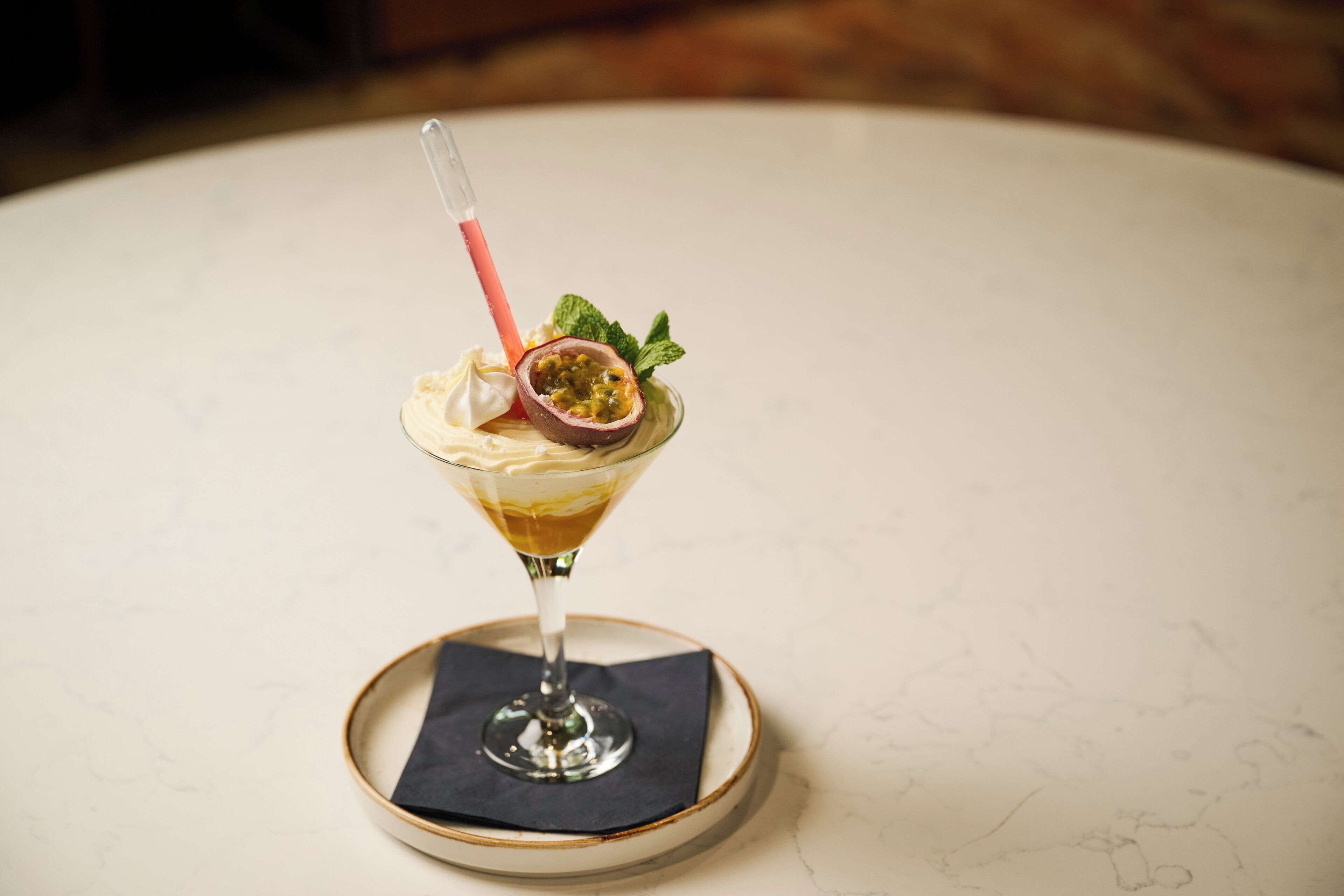 A martini glass with a cocktail, a slice of passion fruit, and mint leaves on a marble table.